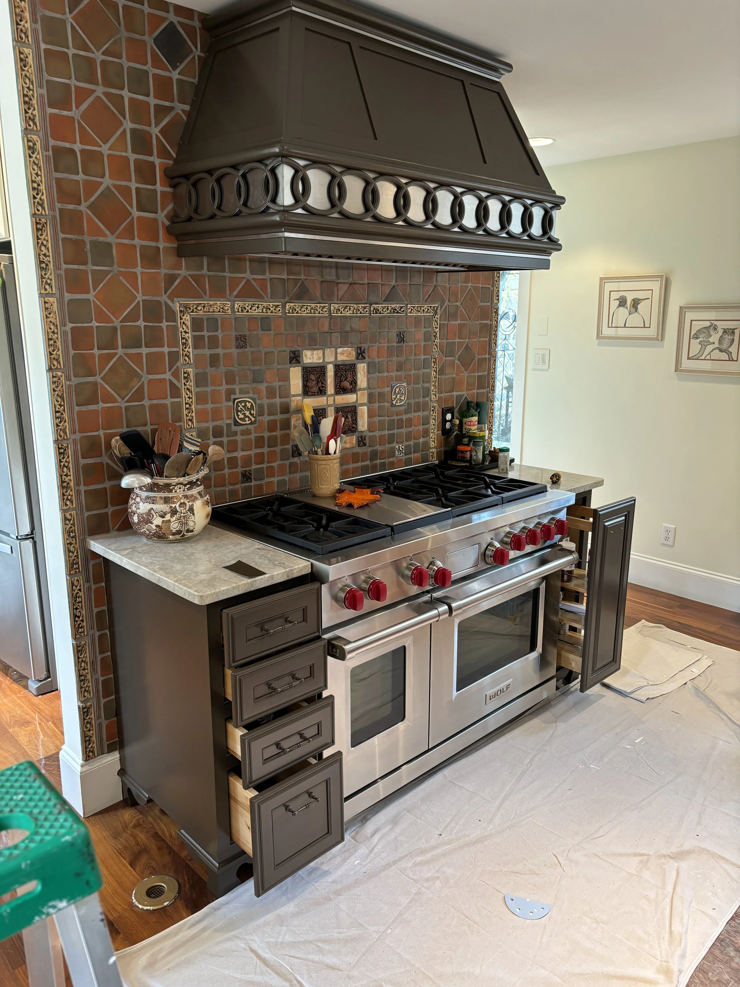 Stainless steel kitchen stove with red knobs, surrounded by brown cabinets and a decorative backsplash with tiles; a large wooden range hood above, and a countertop with utensils and bottles to the side.