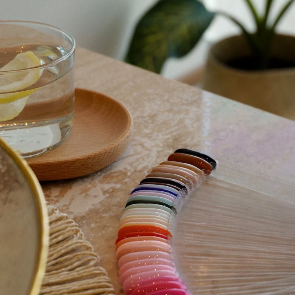 A wooden palette with various shades of nail polish samples, a glass of water with lemon, and a small wooden dish on a beige countertop, with a potted plant in the background..