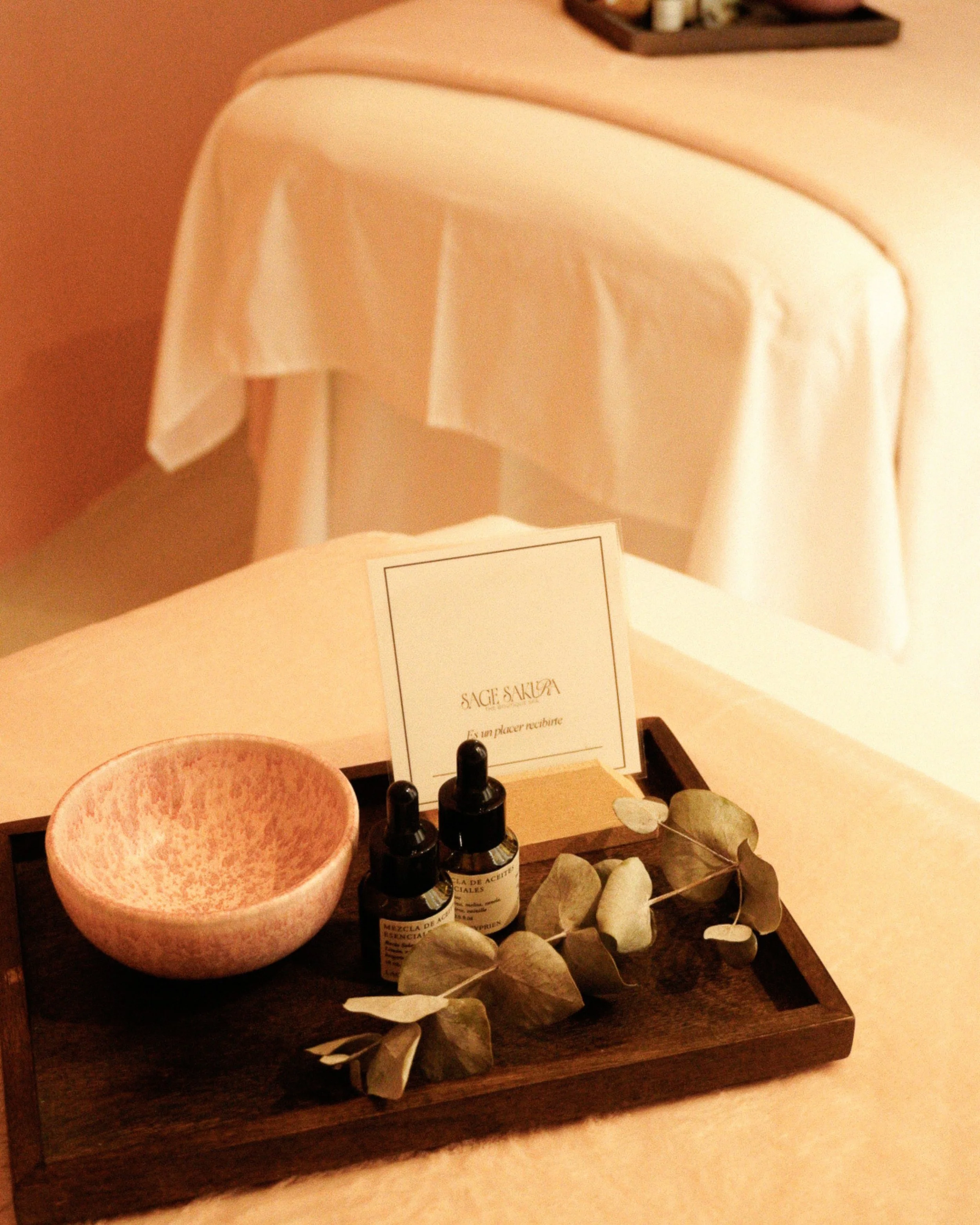 A cozy massage room with warm lighting, wooden accents, and a massage table in the center. The ceiling has decorative lights resembling starry skies. Shelves with products and decor are at the back, with robes hanging on the side.