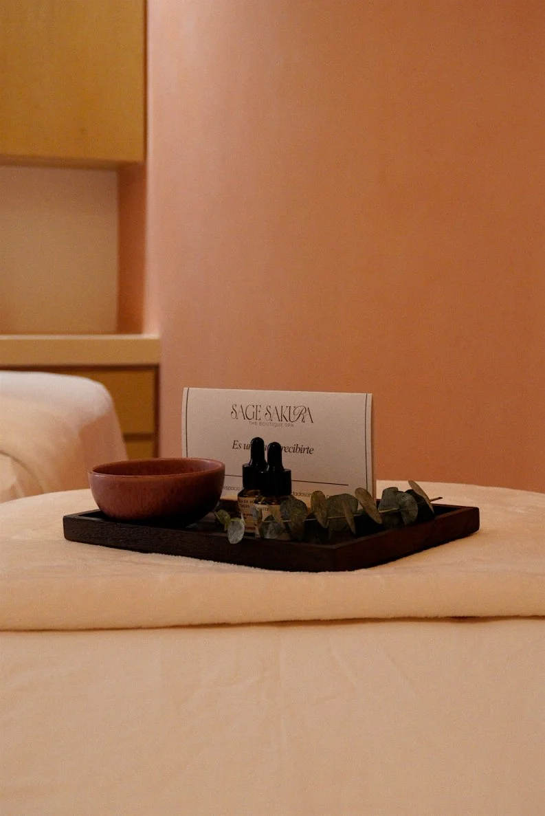 A cozy massage room with warm lighting, wooden accents, and a massage table in the center. The ceiling has decorative lights resembling starry skies. Shelves with products and decor are at the back, with robes hanging on the side.