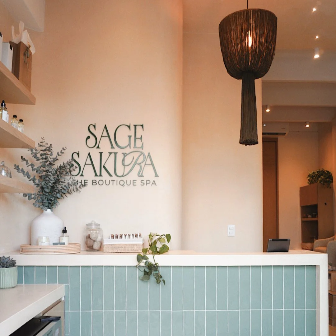 Reception area of Sage Sakura boutique spa with a green tiled front desk, computer, decorative plant, and minimalist decor.