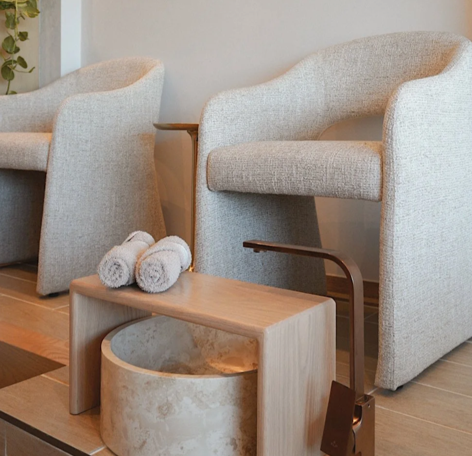 A modern, minimalist interior with a marble-topped table, wire chairs, and a built-in shelf filled with small colorful bottles, all in neutral tones and warm lighting.