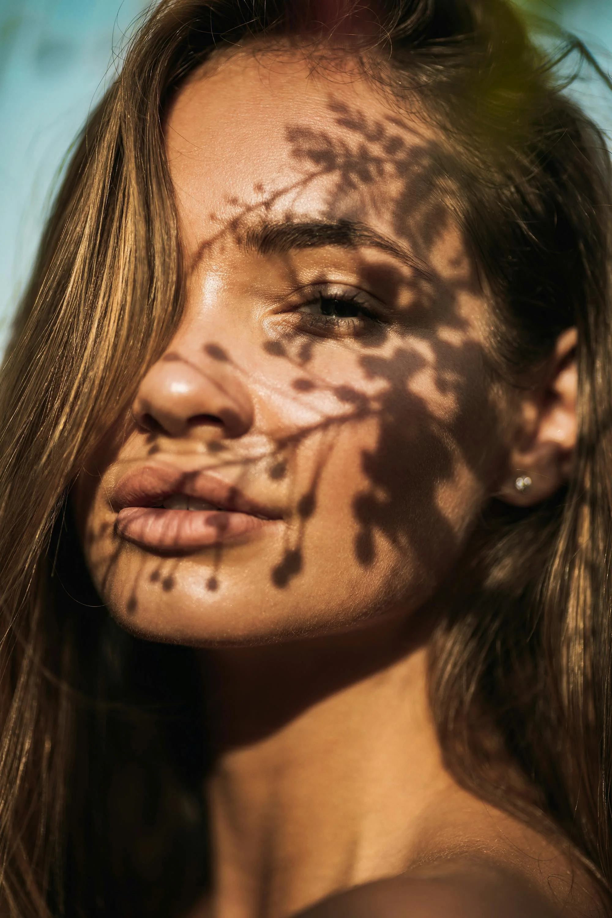 Close-up of a woman's face with sunlight creating shadow patterns of leaves on her forehead and cheek. She has long brown hair, closed eyes, and a peaceful expression.