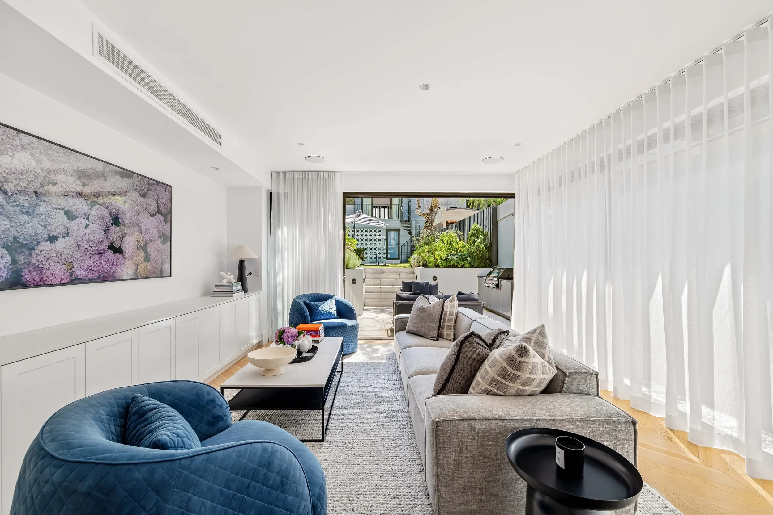 Modern living room with light-colored sofa, blue armchairs, large window with white curtains, and outdoor patio in the background.
