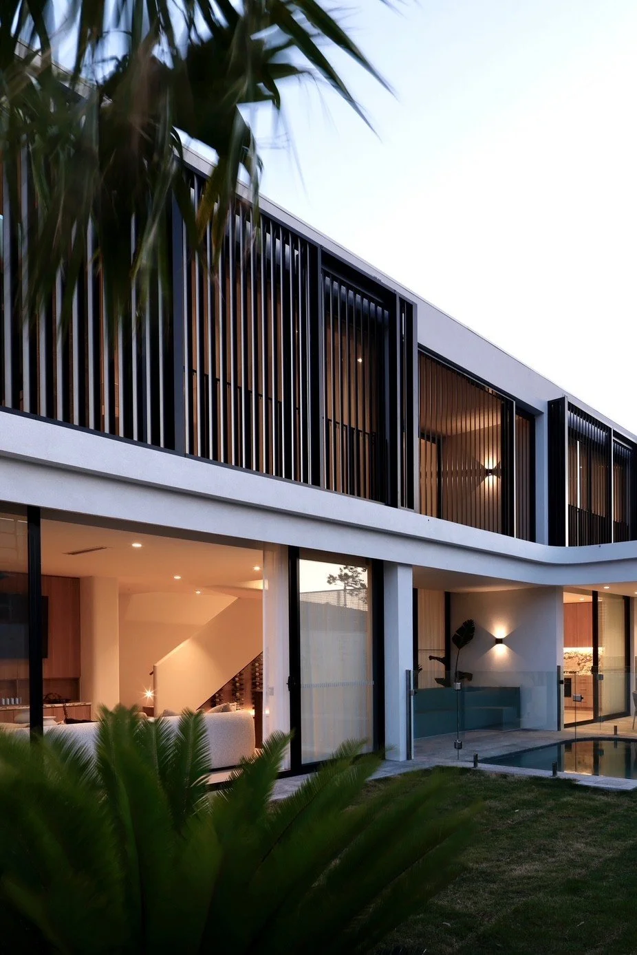 Modern two-story house with large glass windows and wooden slats, illuminated from inside, with a garden and pool area.