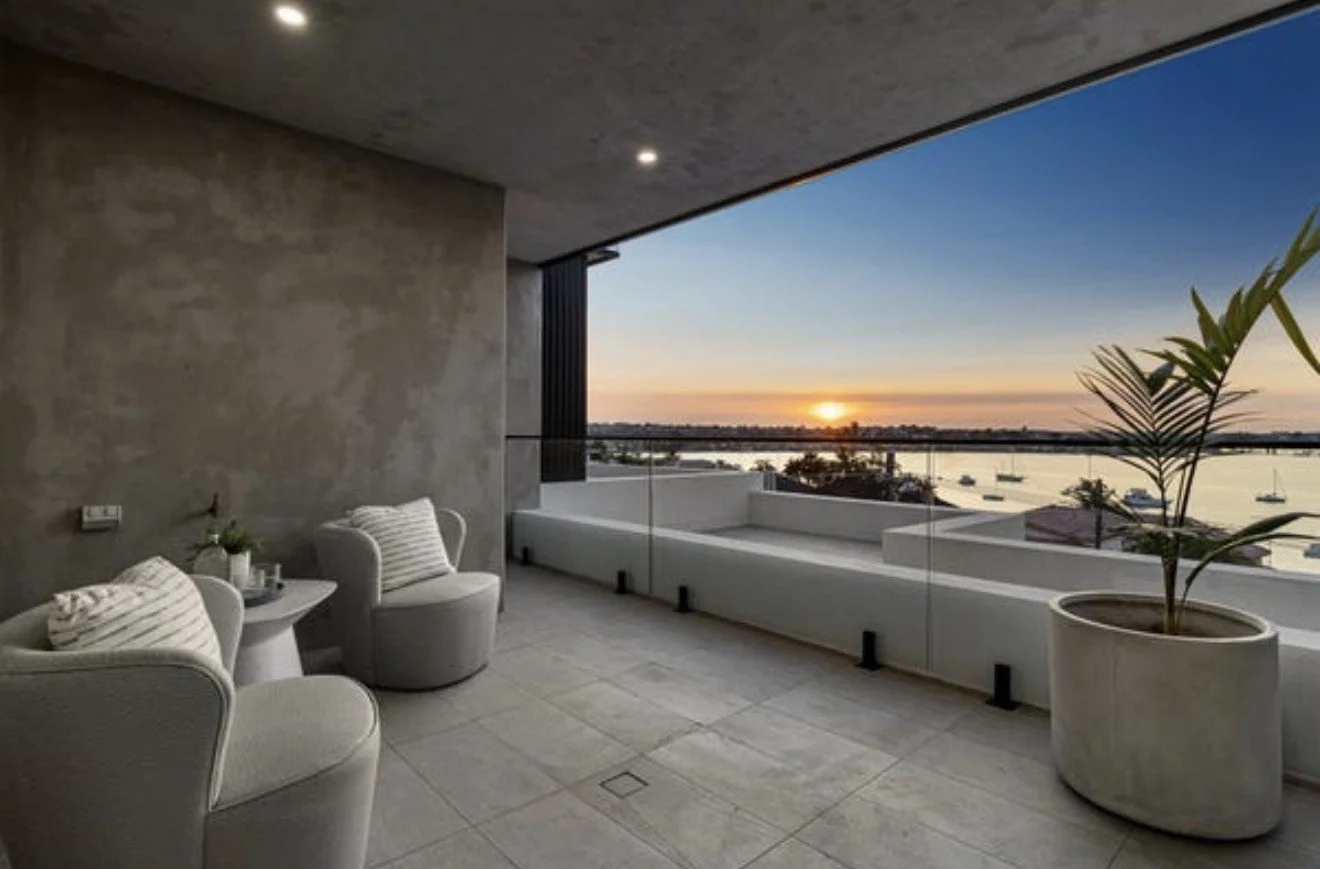 Modern balcony with two white chairs, a small white side table, a potted plant, overlooking a body of water at sunset.