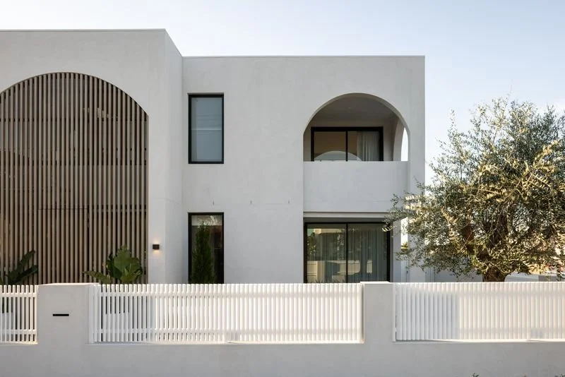 Modern white house with minimalist architecture, featuring a curved wooden slat facade on the left, arched balcony, large windows, and a white picket fence, with a tree on the right side.