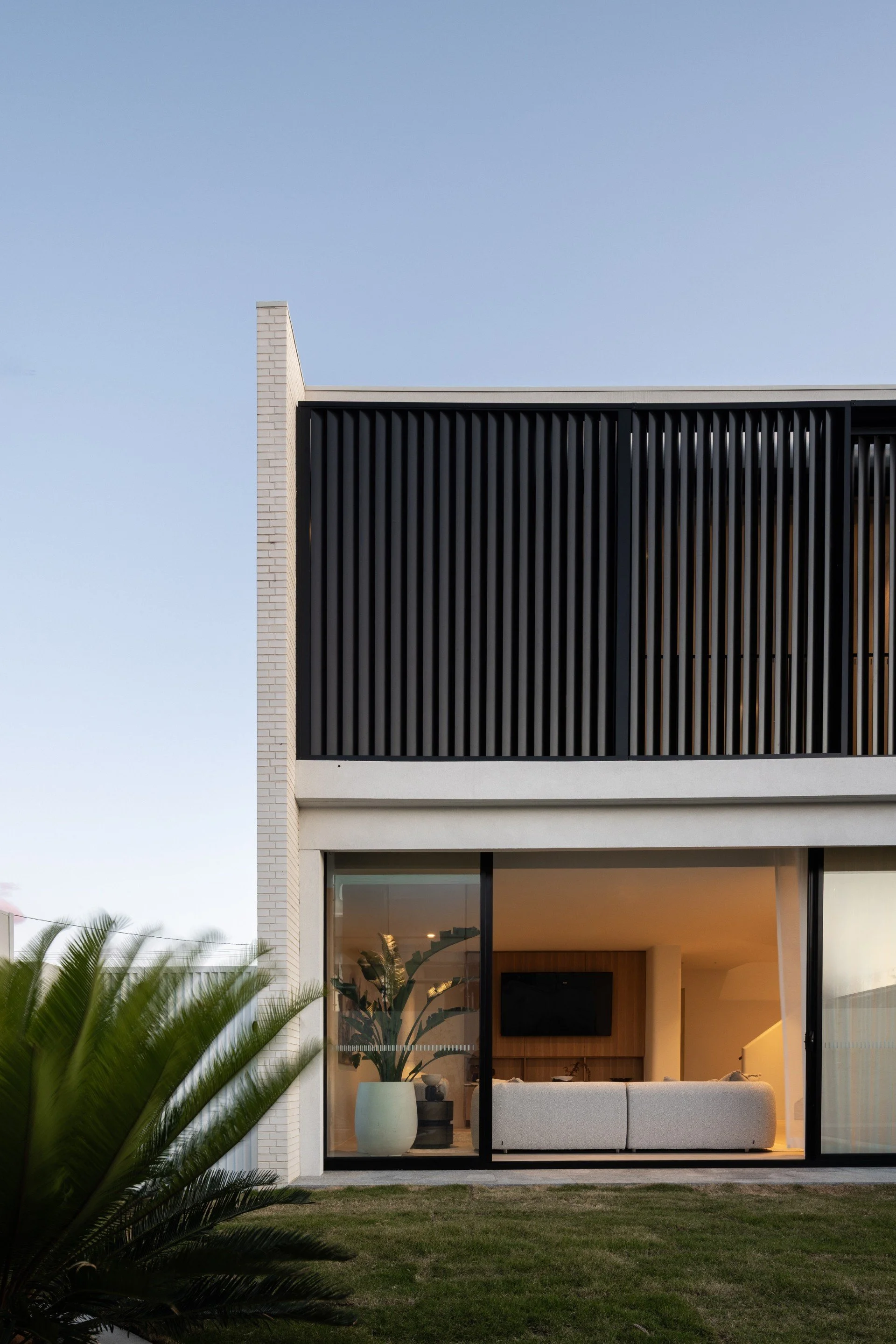 Modern two-story house with a white brick exterior and large glass sliding doors leading to a living room. The upper floor features black vertical slats for a balcony or privacy screen. A large potted plant is visible through the glass, and there is a well-maintained lawn in front.