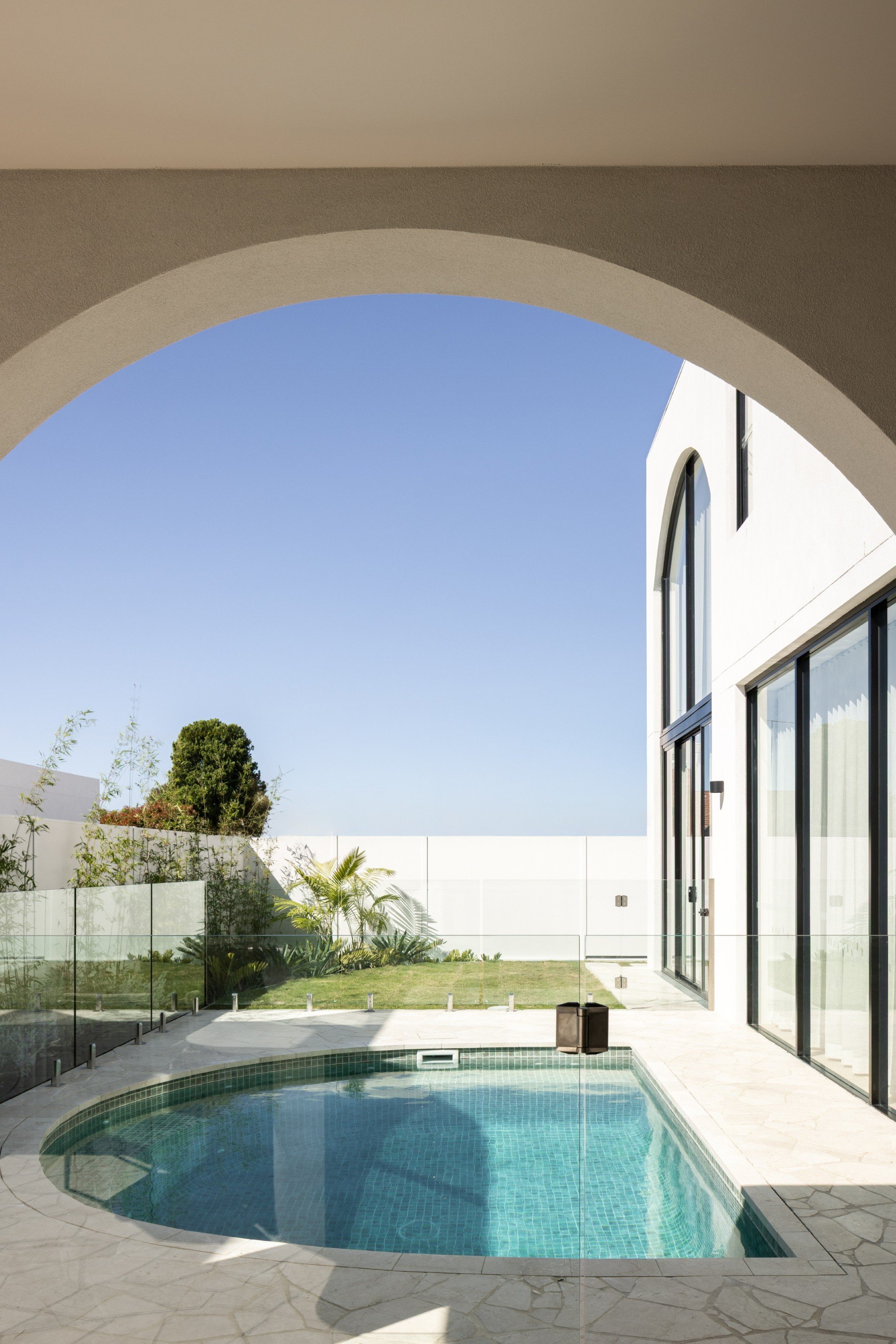 view of a small backyard pool through an arched outdoor structure, with modern white house and blue sky in the background.