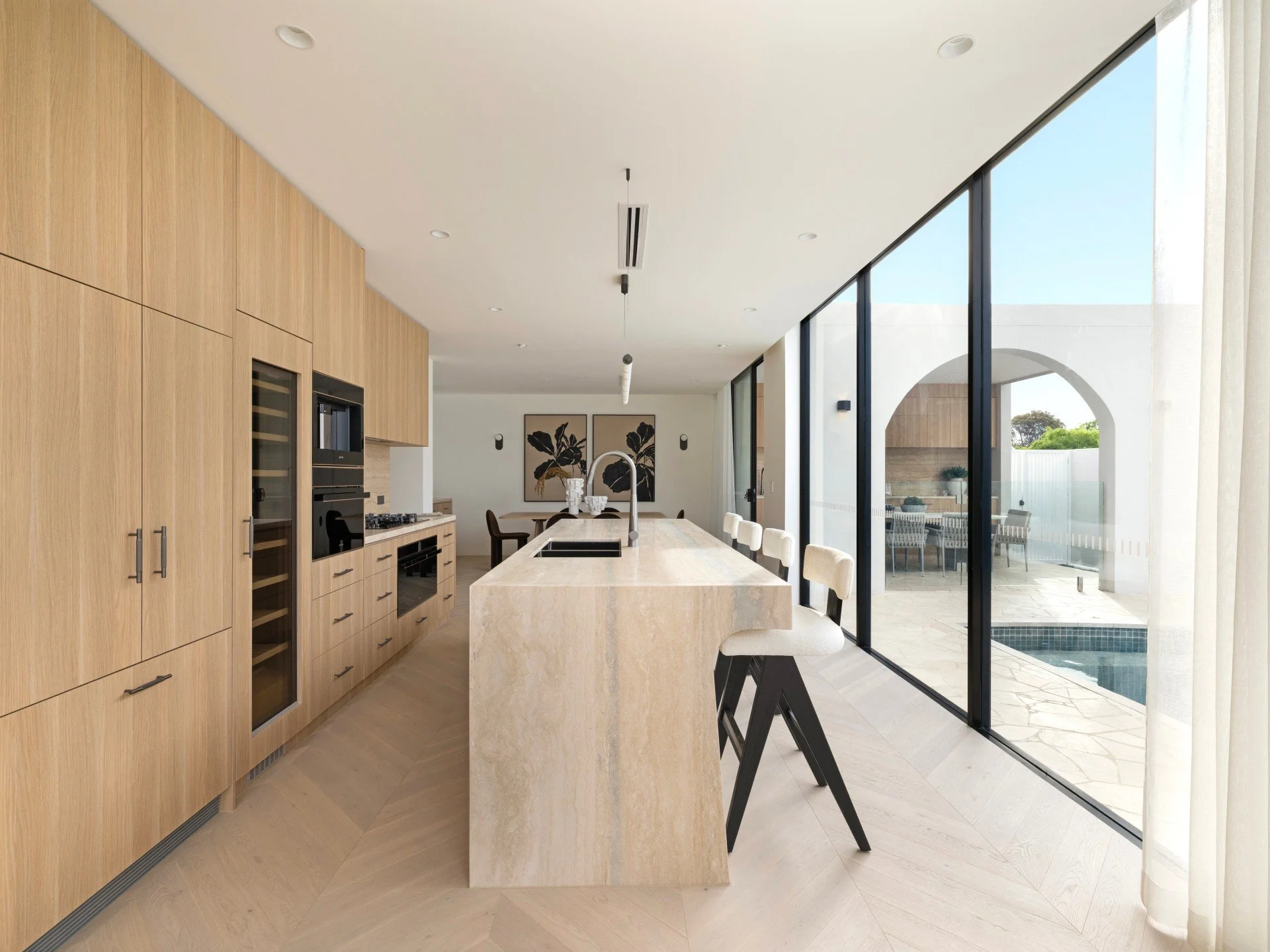 Modern open-concept kitchen with wooden cabinets, light-colored island, bar stools, large glass sliding doors overlooking a patio with a pool, and minimal decor.