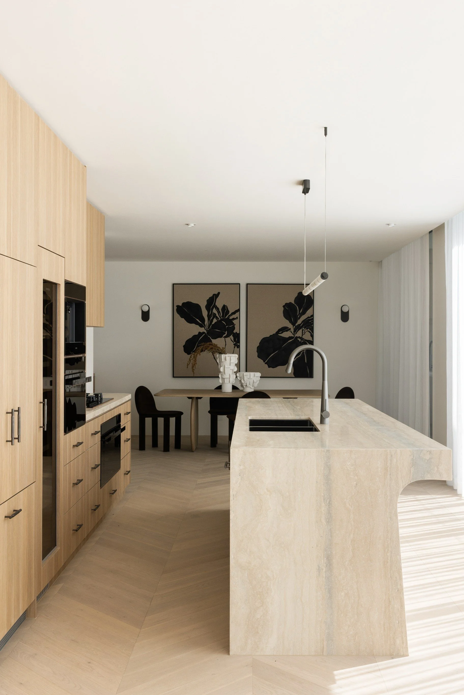 Modern open-plan kitchen with light wood cabinetry, beige marble island with sink, black faucet, and two wall-mounted black lights, leading into a dining area with black chairs and two large black-and-brown artworks on the wall.