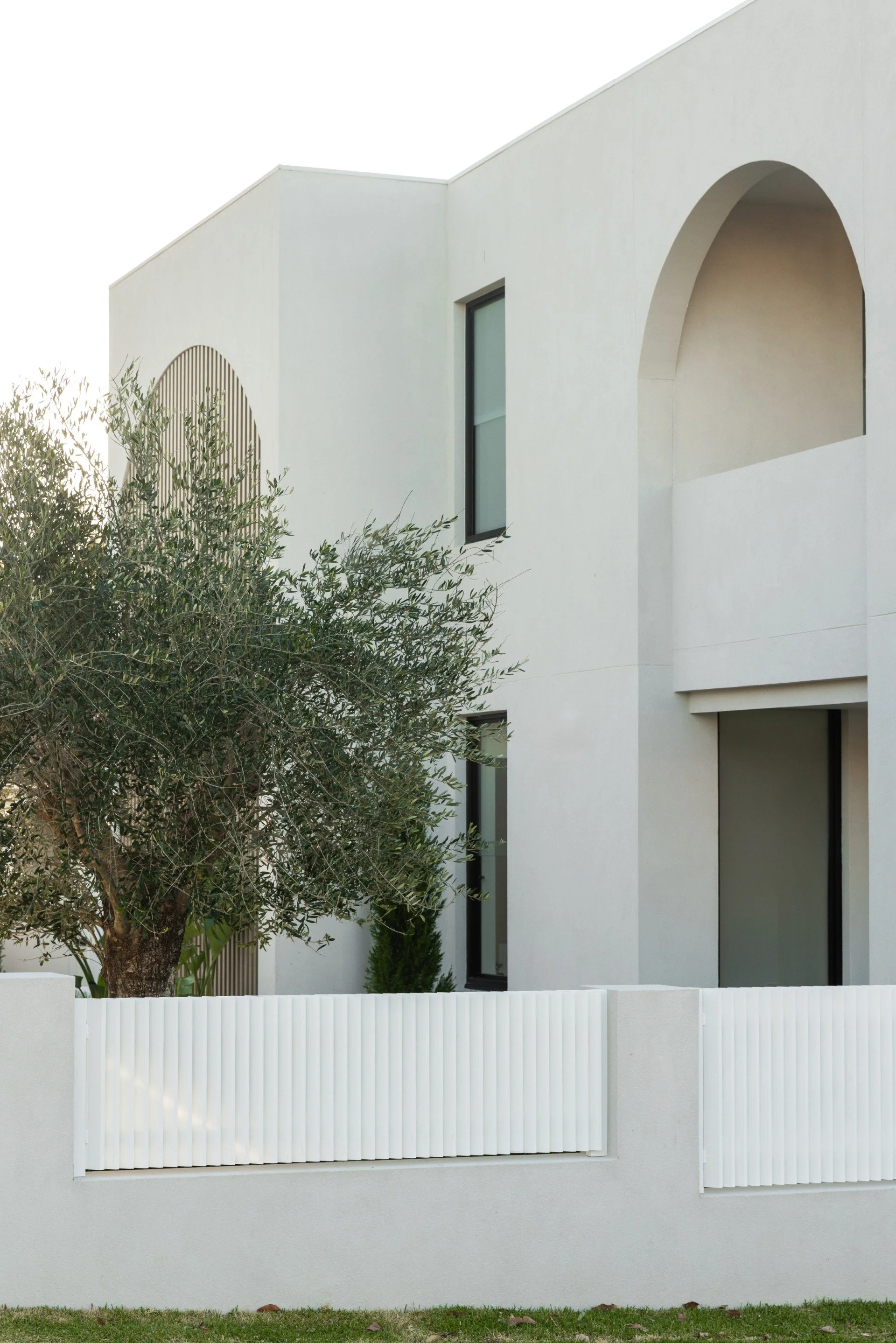 Modern white house with arched and rectangular windows, surrounded by a white fence and a tree in the yard.