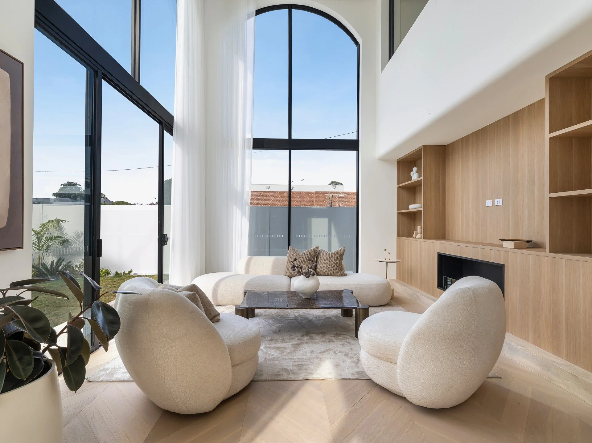 Modern living room with large floor-to-ceiling windows, cream-colored furniture, and wooden wall shelving.