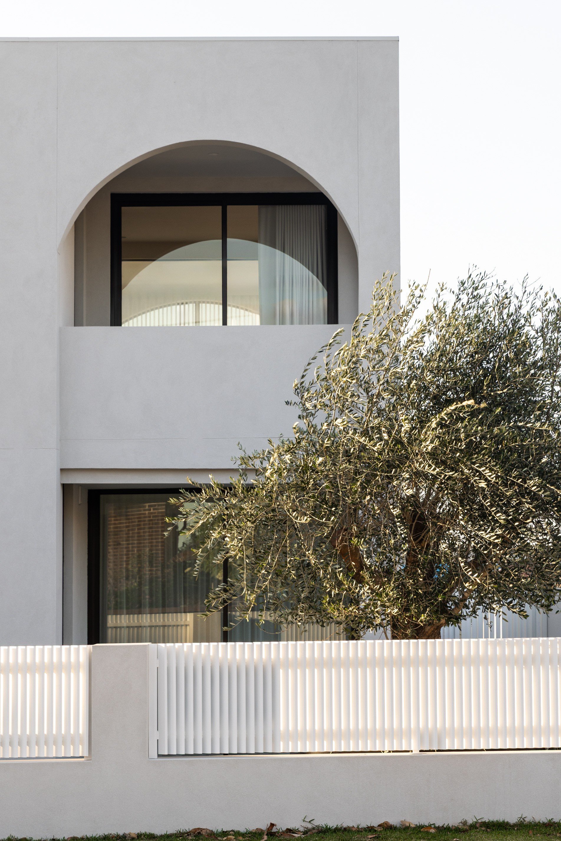 Modern white building with arched window, sliding glass door, and a white fence in front; tree with green foliage outside.