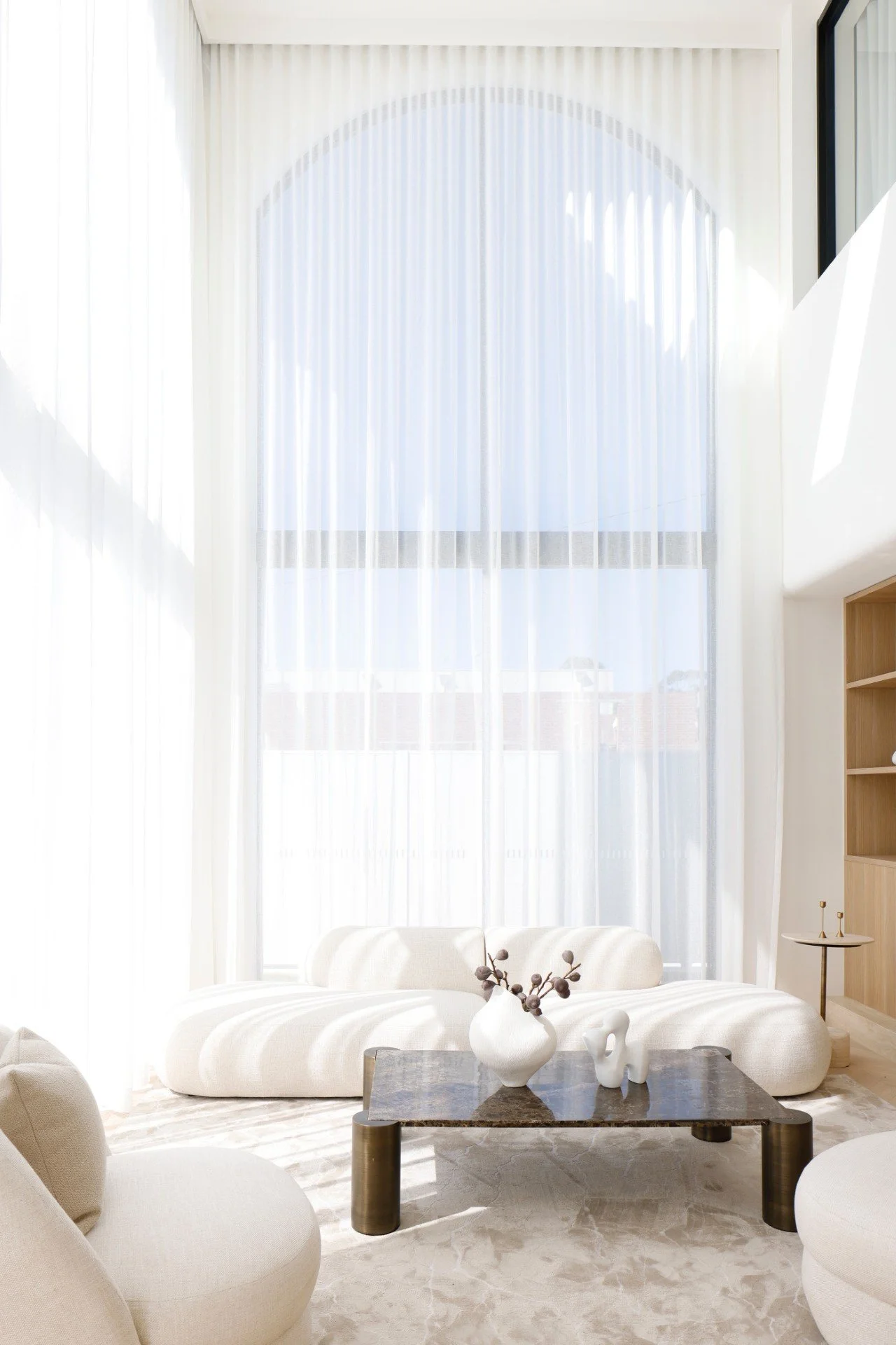 Bright living room with large arched window and sheer white curtains, minimalist white sofa and armchair, marble coffee table with vase and sculptures, neutral color palette.