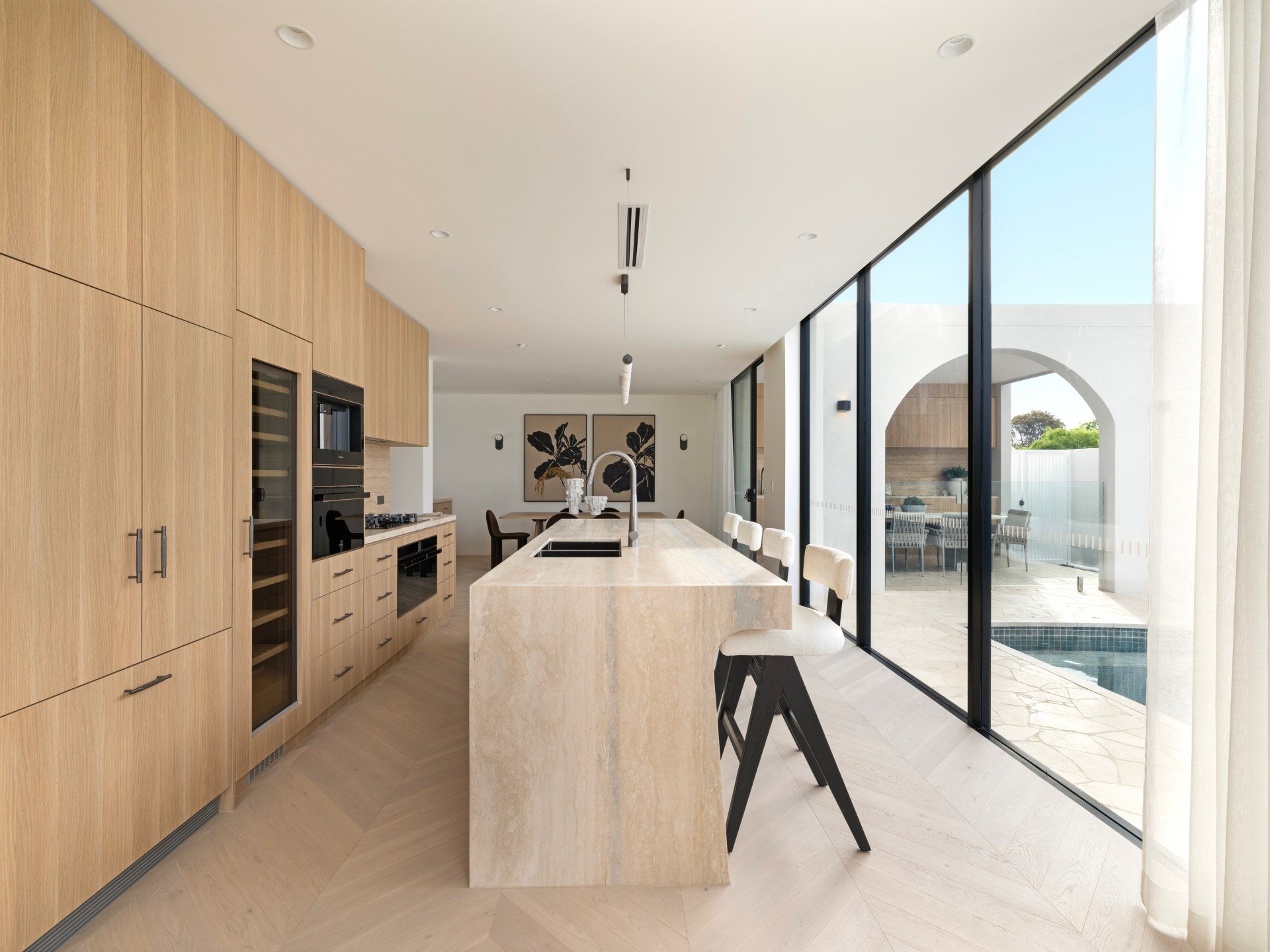 Modern kitchen with light wood cabinets, a large marble island, black and white bar stools, and sliding glass doors leading to an outdoor patio and pool.