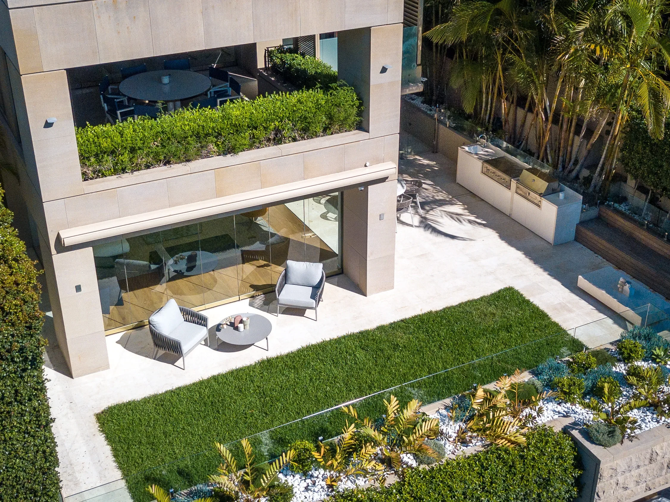Aerial view of a modern backyard patio with outdoor seating, a glass sliding door, a built-in outdoor kitchen with grill, lush green lawn, and tropical plants along the perimeter.
