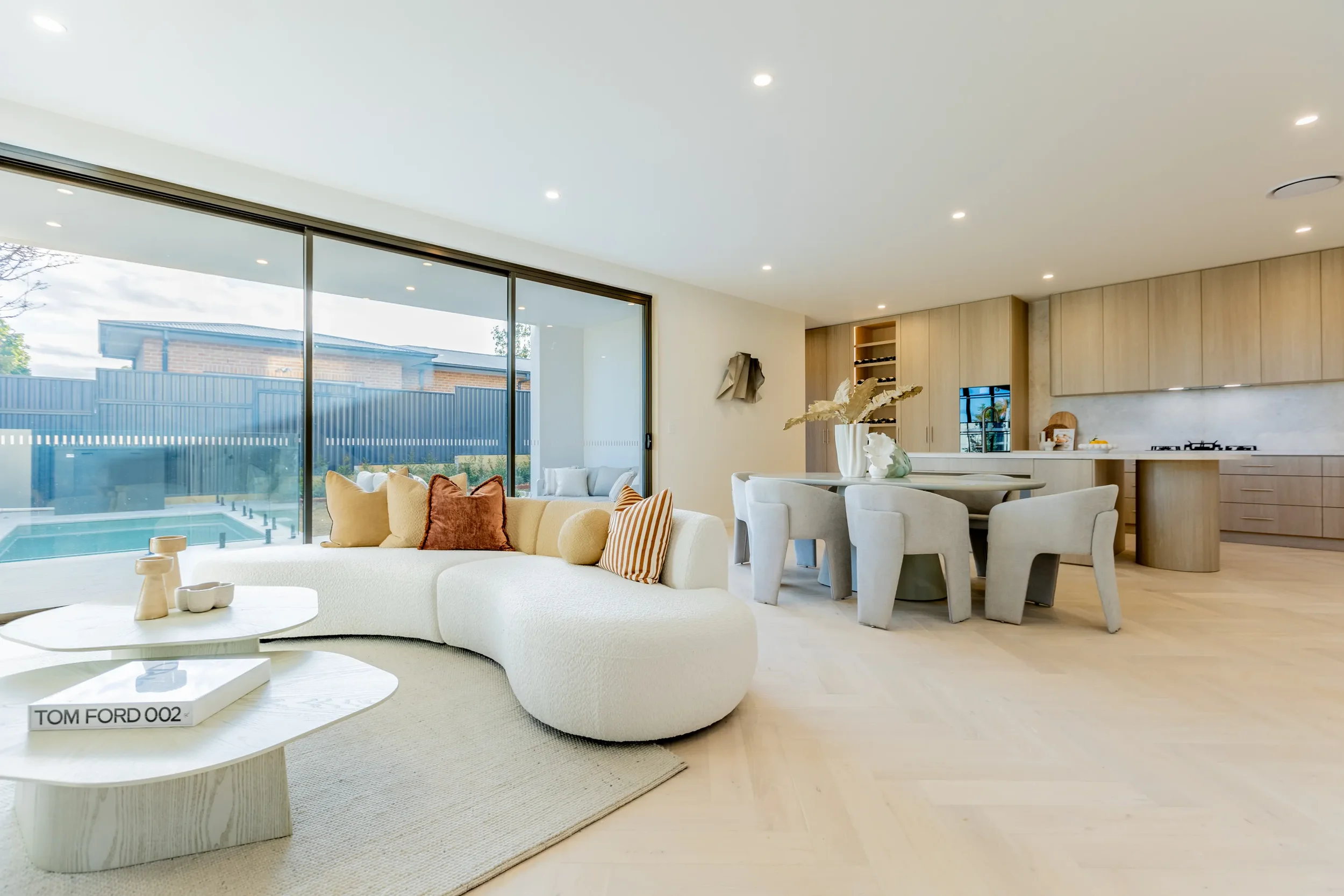 Modern open-concept living room and kitchen with large glass sliding doors, white curved sofa with throw pillows, round white coffee table, dining table with six chairs, and wooden kitchen cabinets.