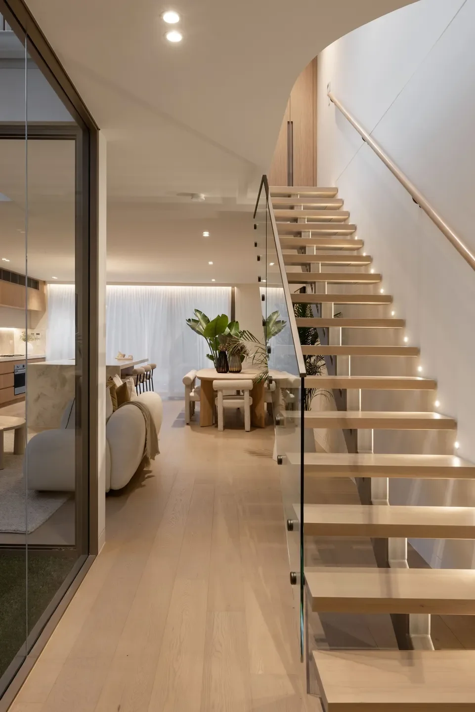 Modern interior view of a home with a wooden staircase, living area, and dining space, decorated with plants and neutral colors.