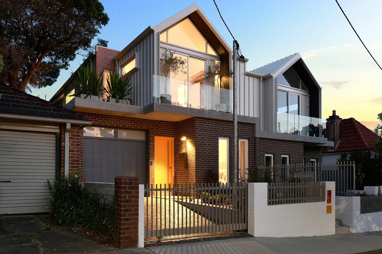 Modern multi-story house featuring brick and metal exterior, large glass windows, and contemporary design elements, illuminated at dusk.