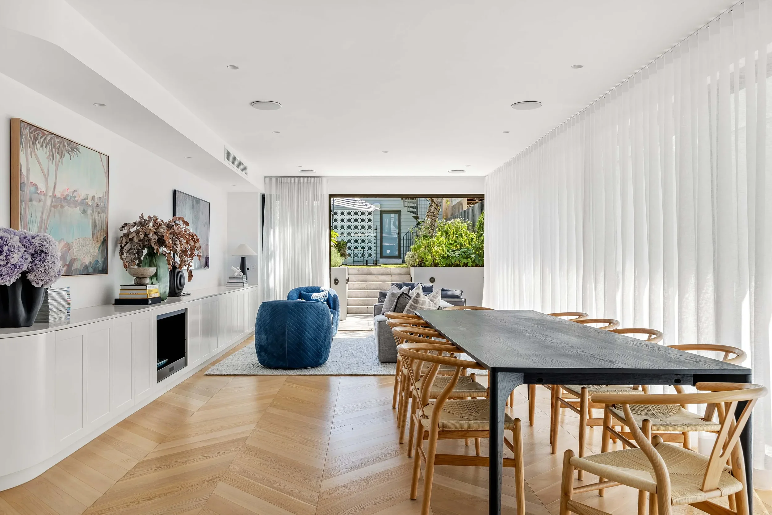 A modern living and dining area with white walls, a large wooden dining table with light wooden chairs, and a sitting area with blue chairs, a sofa, and artwork on the wall, with large windows and sliding glass door leading outside, decorated with plants.