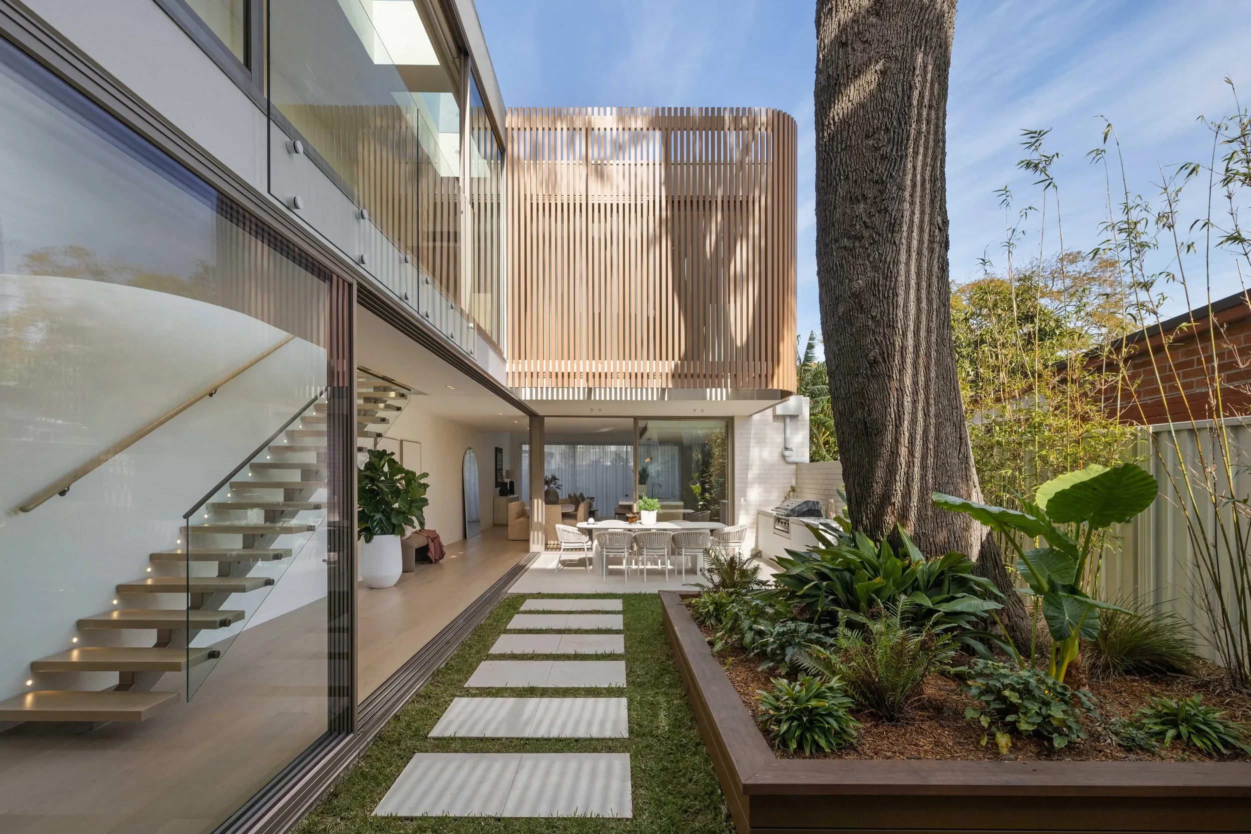 Modern backyard with patio, outdoor dining area, large tree, and landscaped garden with lush plants