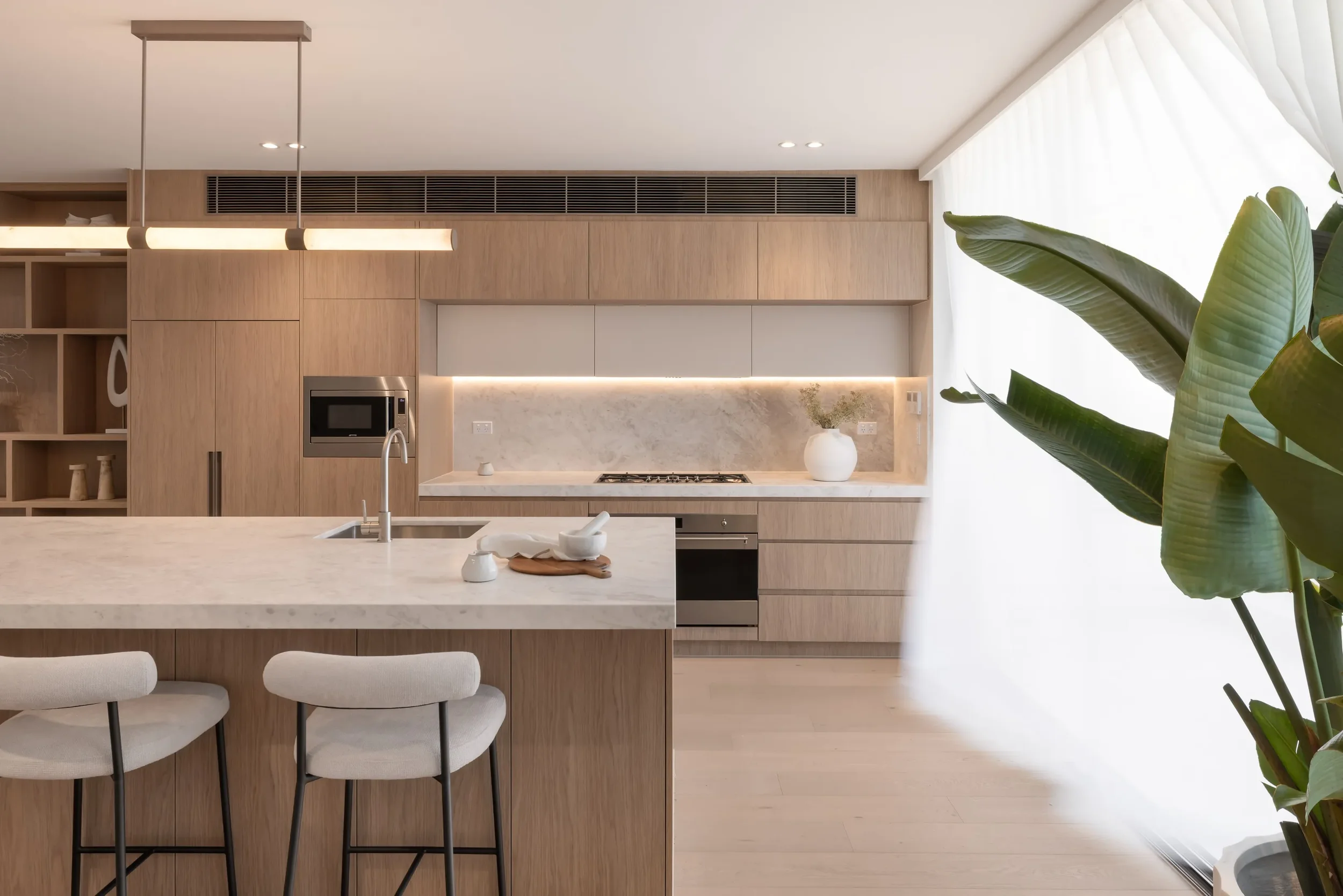 Modern kitchen with light wood cabinetry, marble backsplash, island with barstools, and large window with sheer curtains, accompanied by green plants.