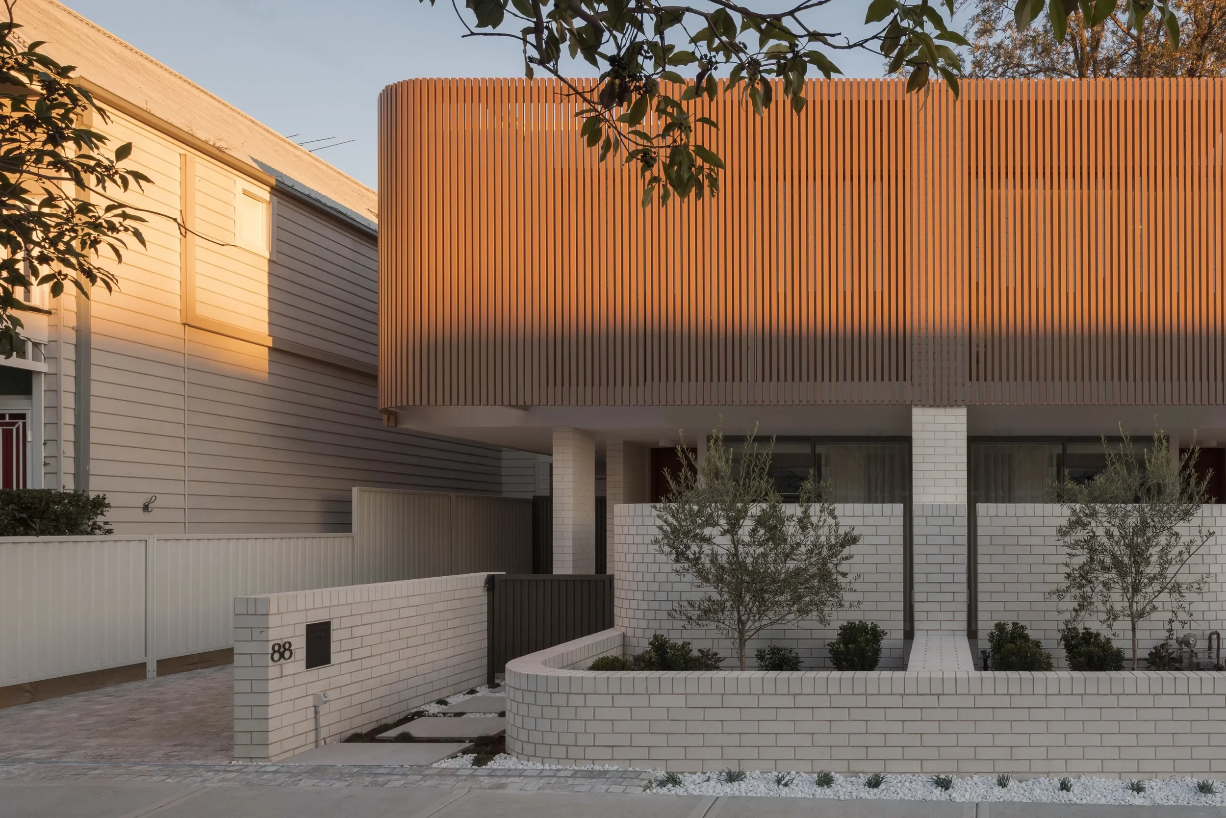 Modern house with white brick walls, wooden slatted upper facade, small trees and landscaped front yard, in warm sunset lighting.