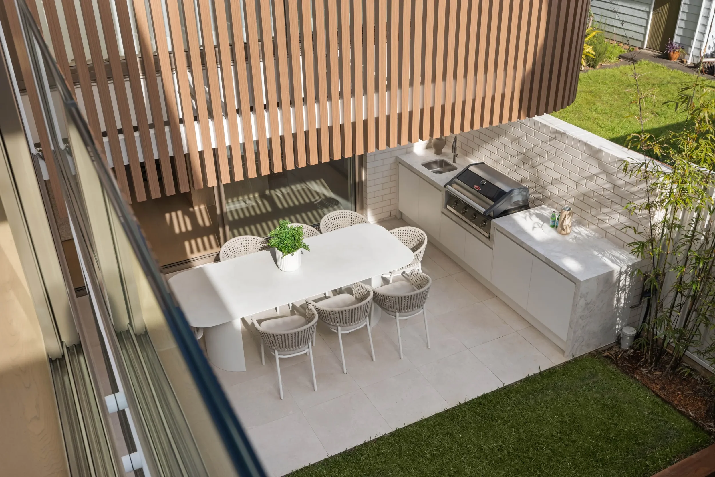 Outdoor patio area with a white dining table and six chairs, a built-in outdoor kitchen with a grill, a small sink, and white cabinets, a potted plant on the table, and a wooden privacy fence with a garden and grass in the background.