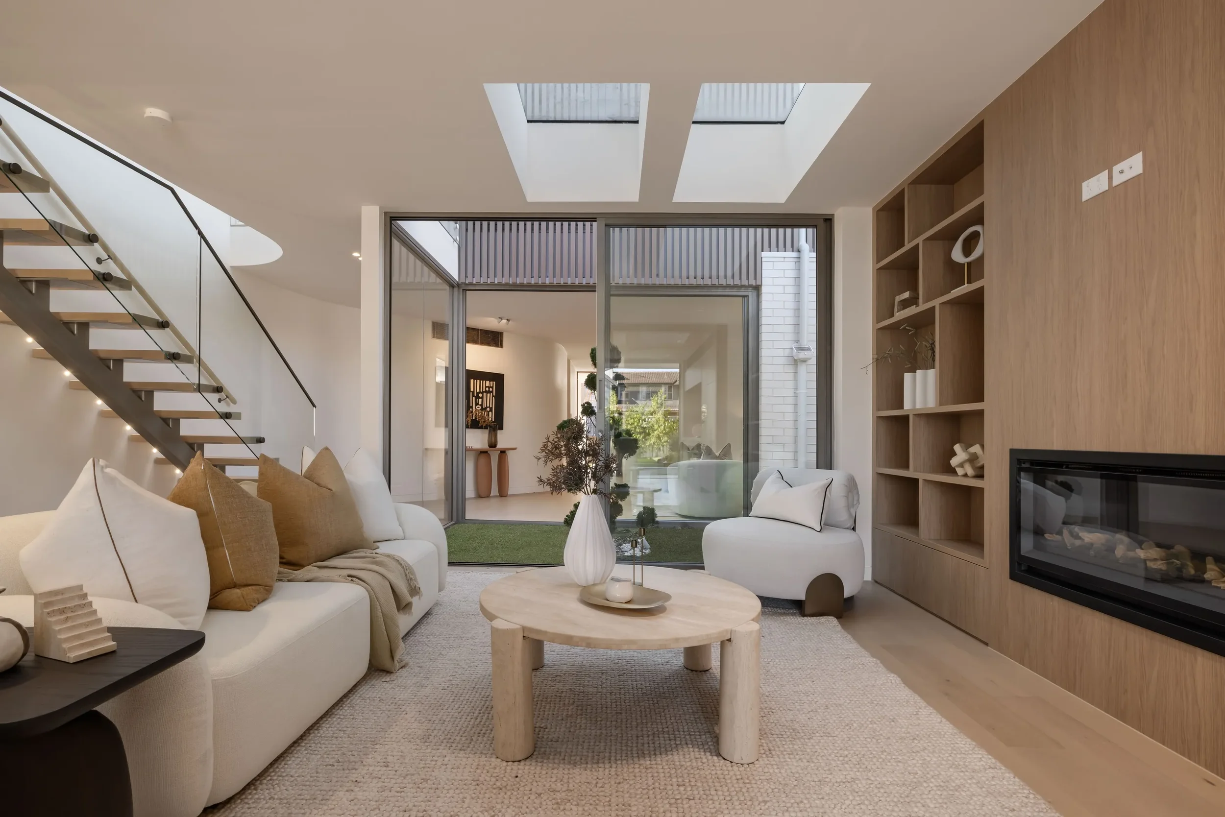 Modern living room with white sofa, armchair, wooden coffee table, built-in wooden shelves, fireplace, and large glass sliding doors leading to an outdoor patio with greenery.