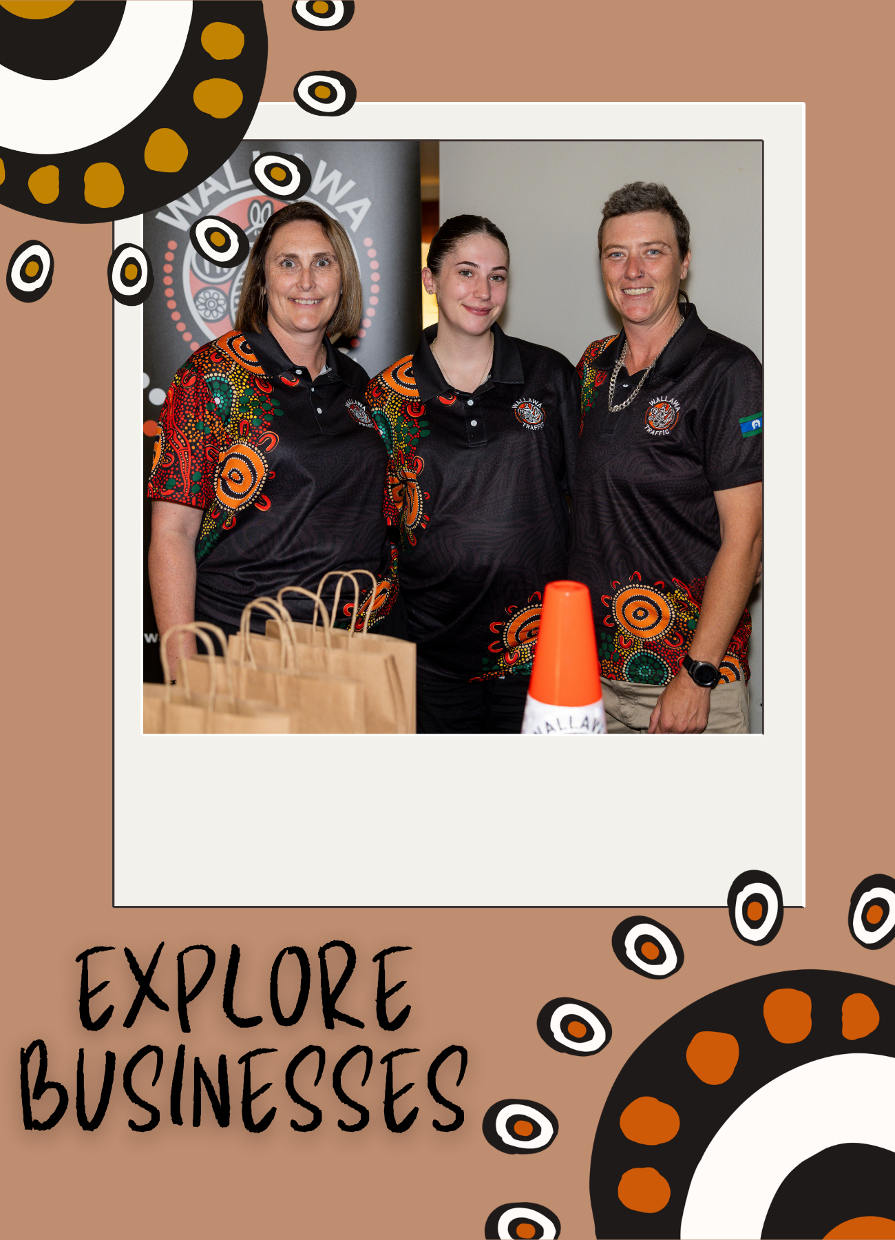 Three women in black shirts with colorful patterns, smiling, standing together at an event. Behind them is a black banner with a logo and text. In front of them are paper bags and an orange traffic cone with white paper.
