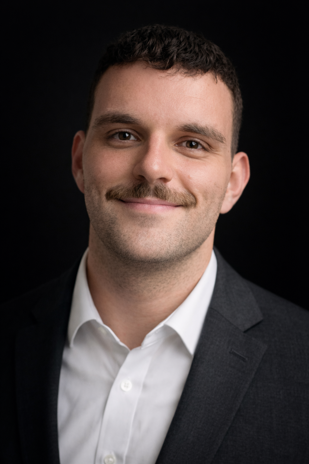 Headshot of a young man with short dark hair, mustache, wearing a white shirt and dark suit, smiling against a black background.