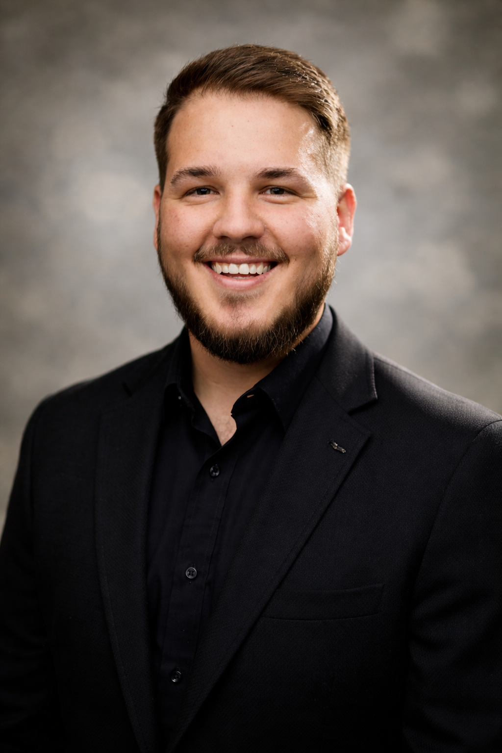 A smiling young man with a beard, dressed in a black suit and black shirt, posing against a gray background.