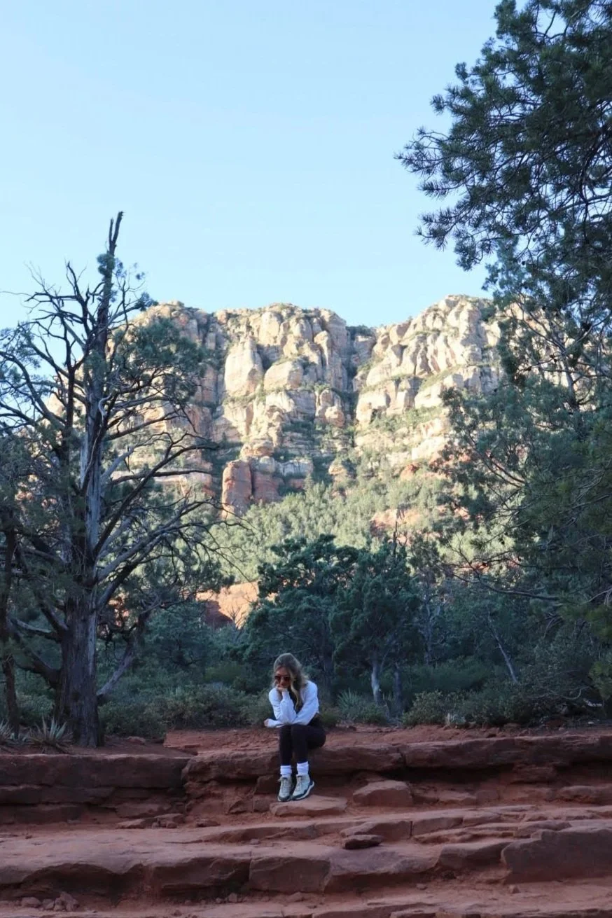 A woman sitting on red rock steps in a natural landscape with trees and cliffs in the background under a clear blue sky.