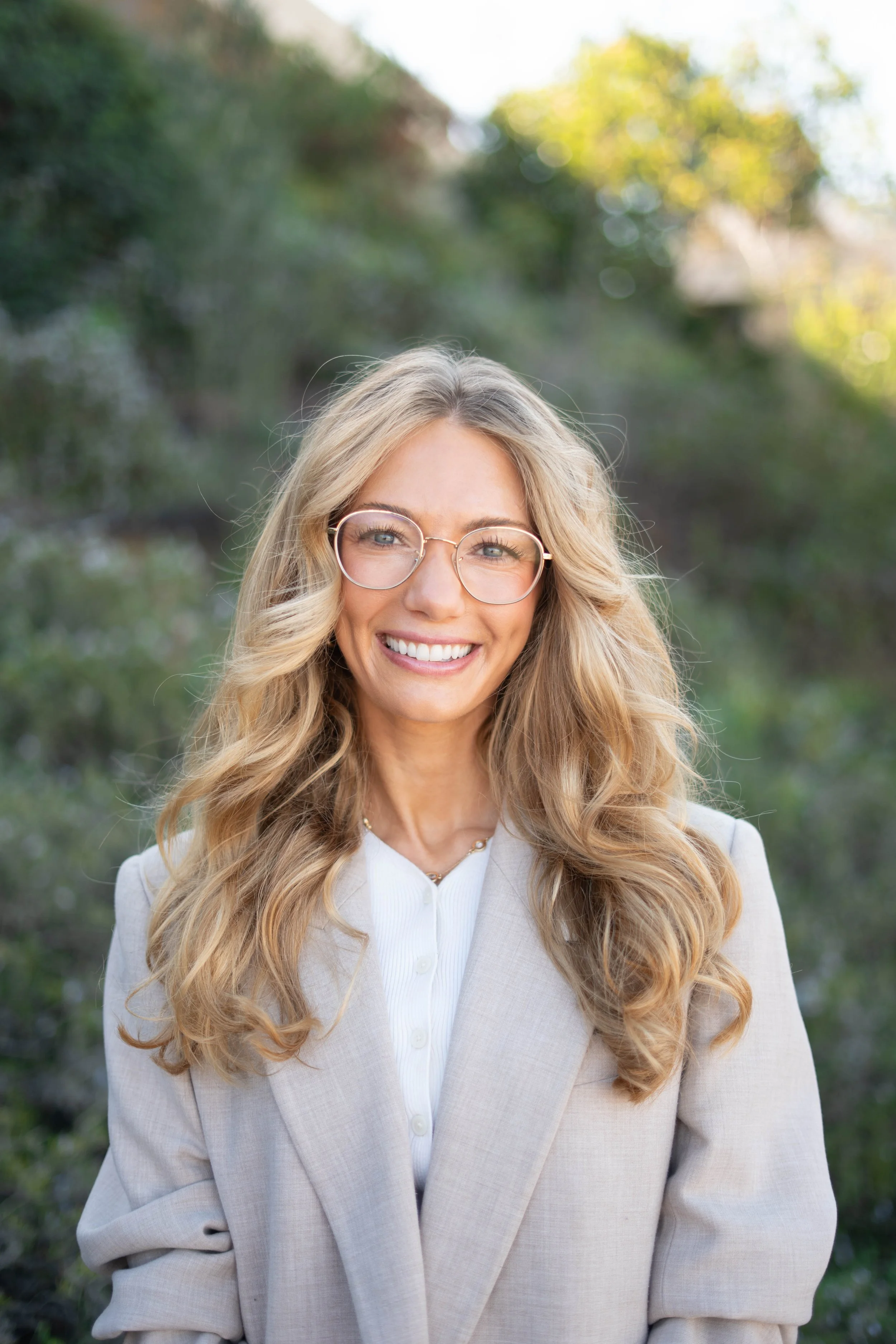 A woman with long blonde hair, wearing glasses and a light-colored blazer, smiling outdoors with a blurred green background.