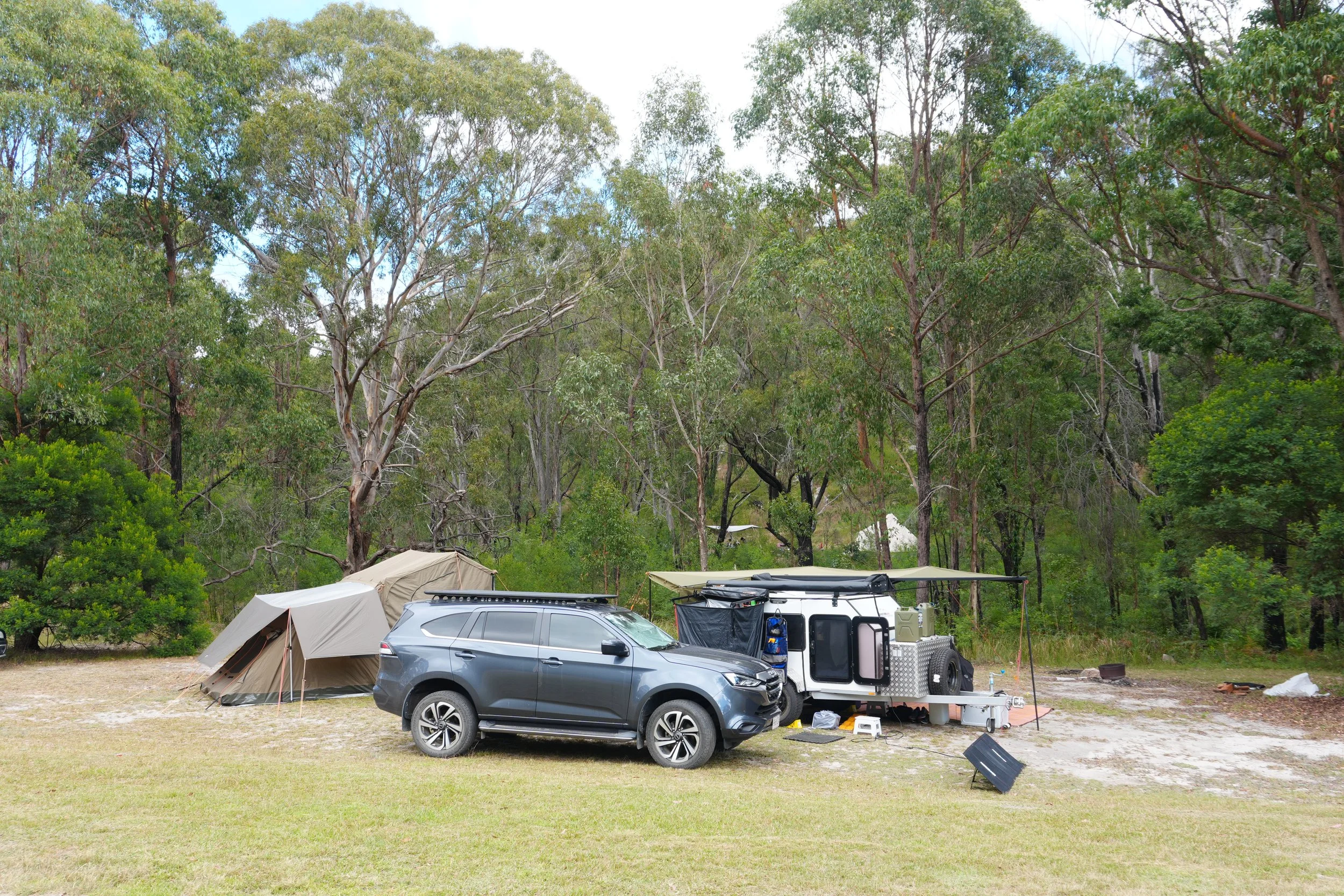 A campsite with tents, a parked gray SUV, and trailer equipment set up on a grassy area surrounded by tall trees under a partly cloudy sky.