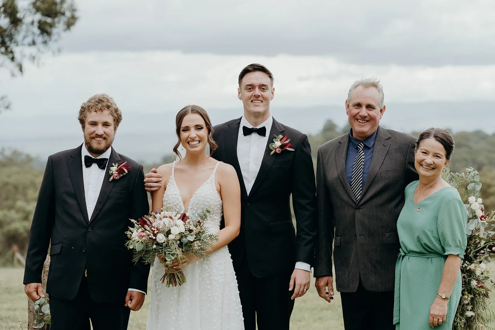 A wedding group portrait outdoors with five people: a bride, groom, and three family members, smiling and dressed in formal wedding attire, standing on a grassy field with trees in the background.