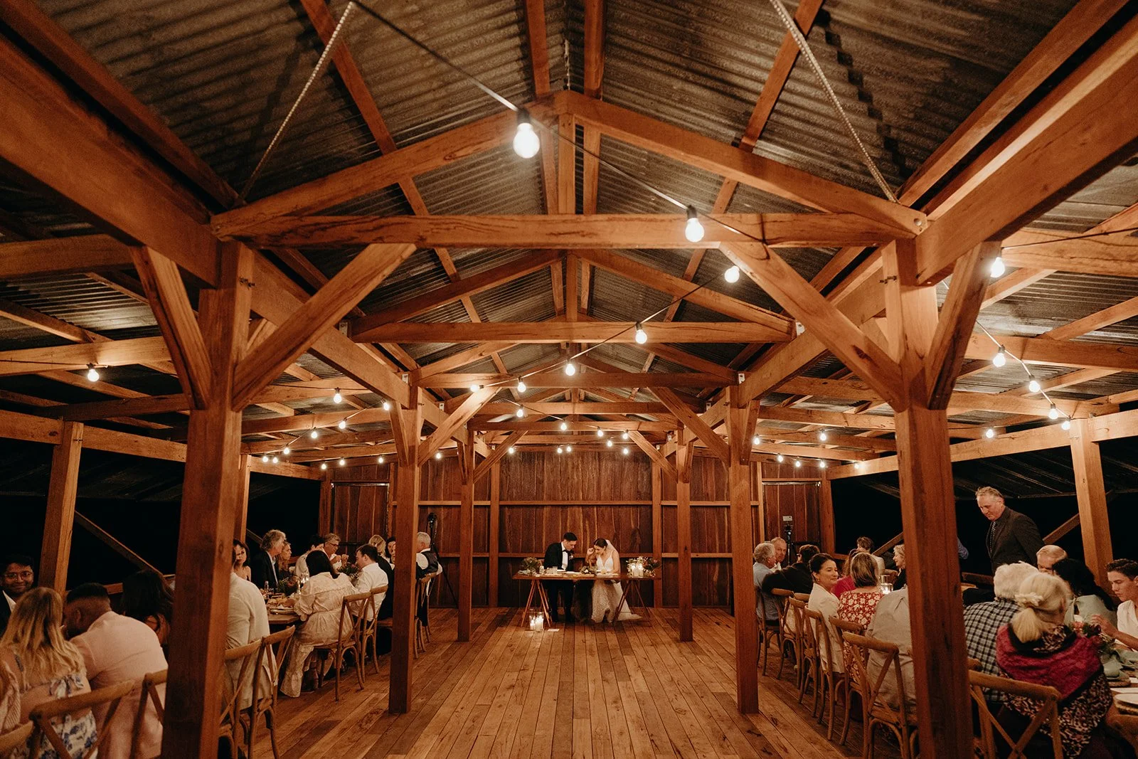 Wooden pavilion wedding reception with string lights, guests seated at tables, and a bride and groom at the head table.