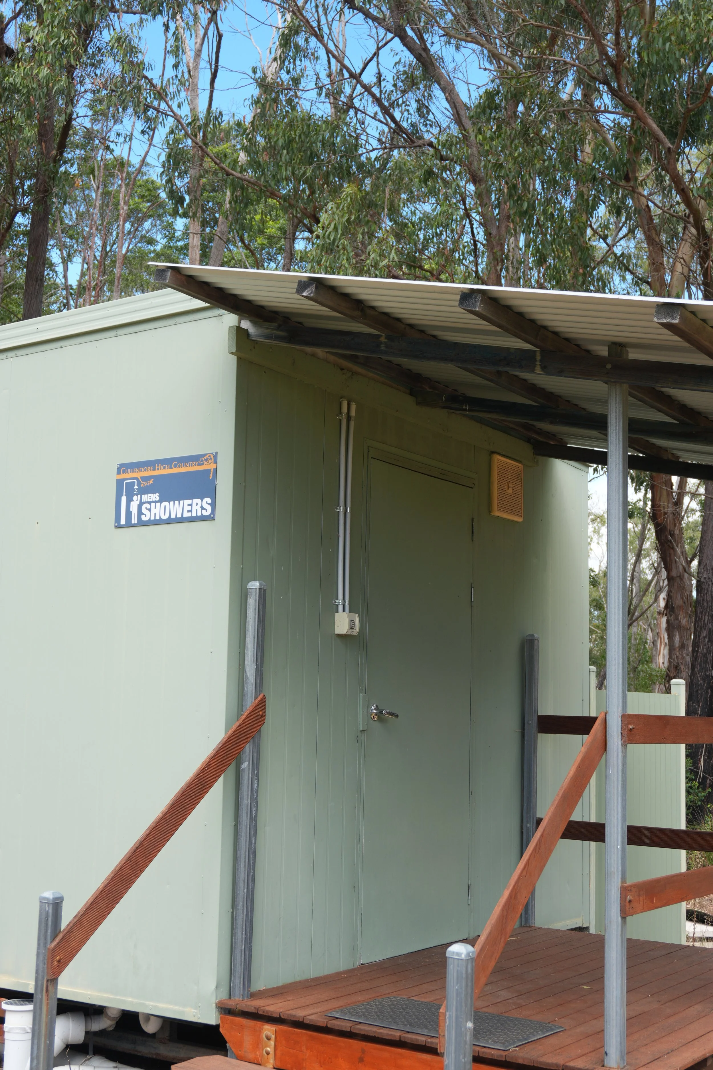 A green outdoor shower building with a sign indicating it is for men's showers, featuring trees and blue sky in the background.