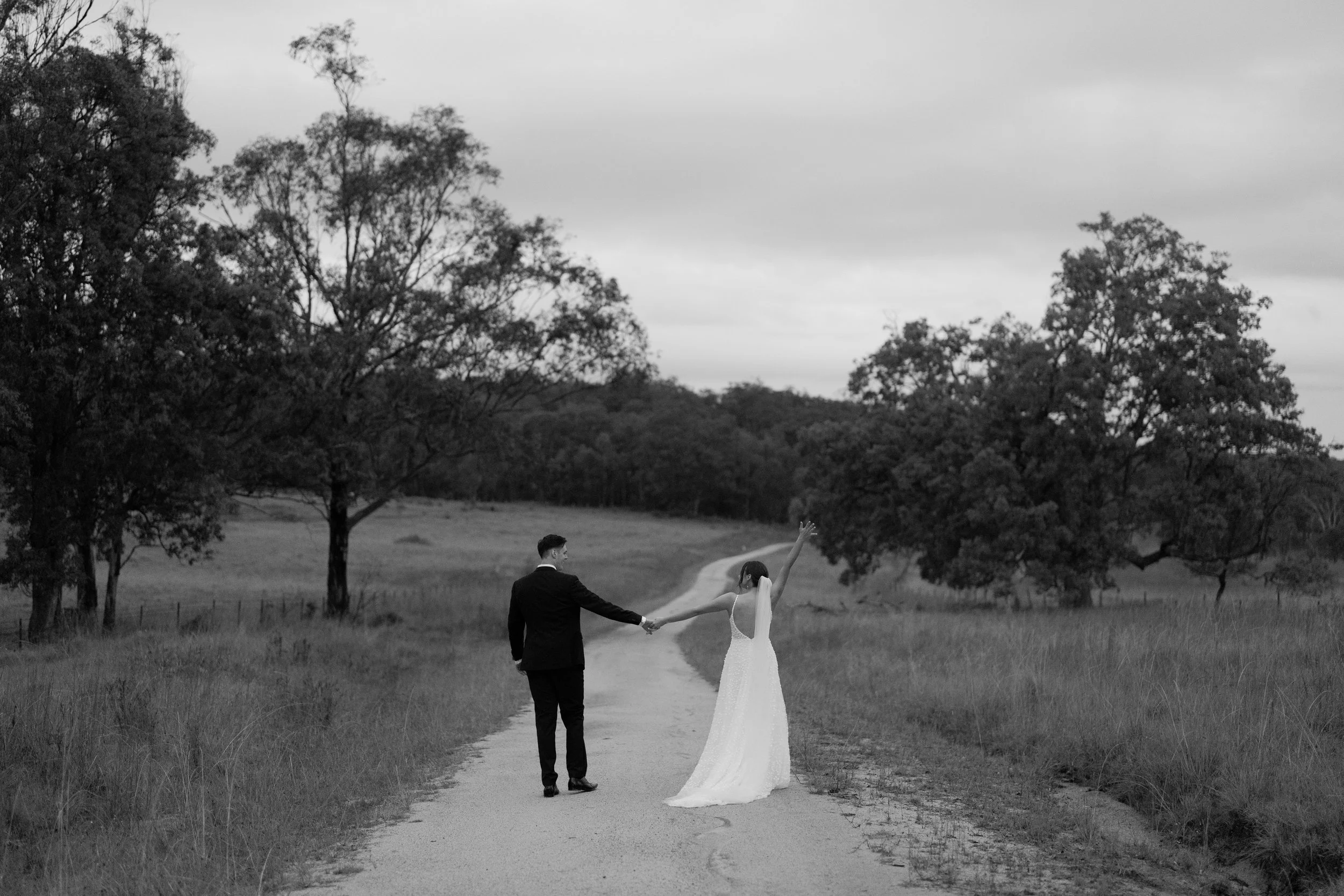 A black and white photo of a couple walking hand in hand on a rural path surrounded by trees, with the woman in a wedding dress and the man in a tuxedo, the woman raising her arm in a joyful gesture.