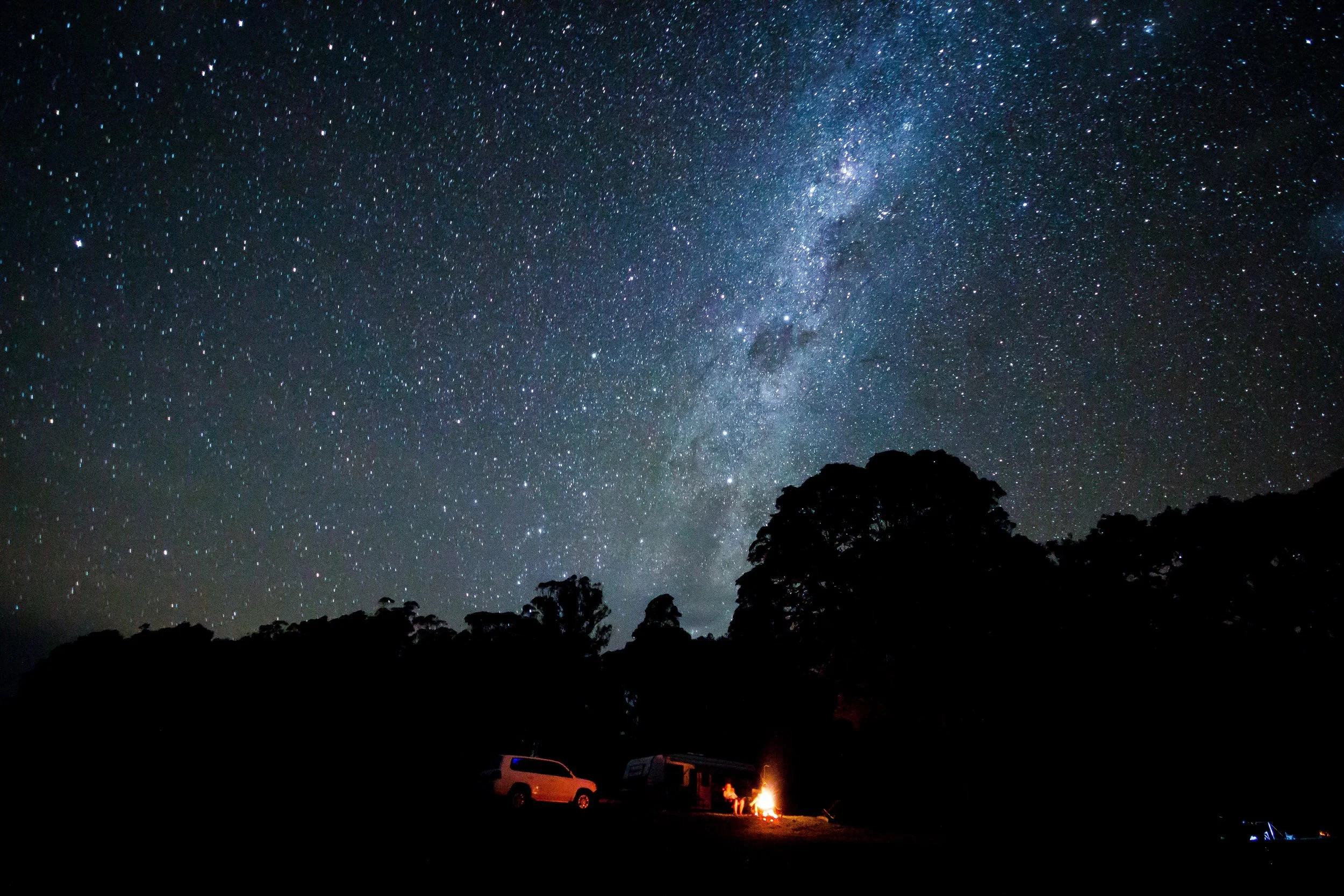 Night sky filled with stars and the Milky Way galaxy. Silhouettes of trees and parked vehicles with a campfire in the foreground.