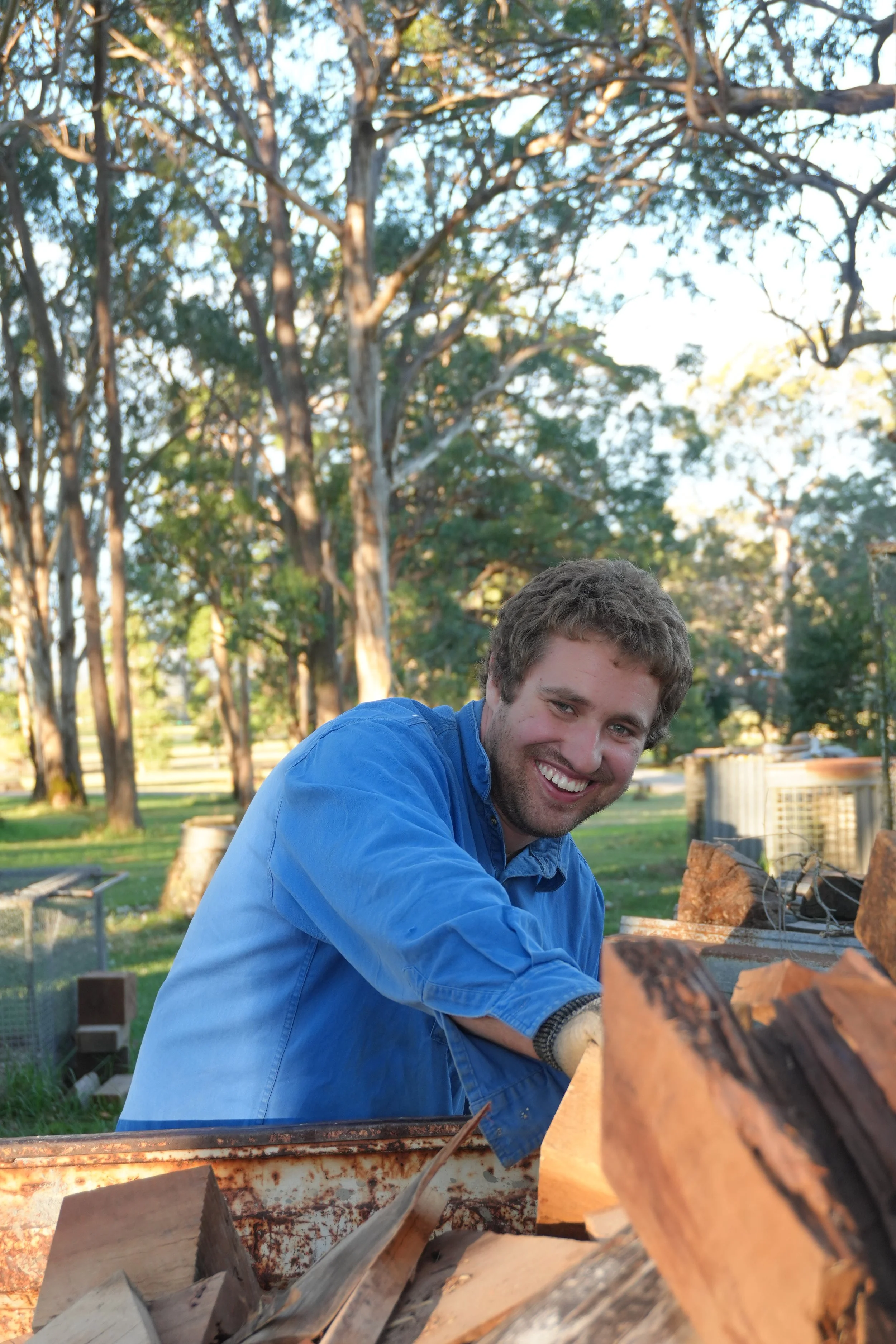 Smiling man in blue shirt chopping wood outdoors with trees in the background