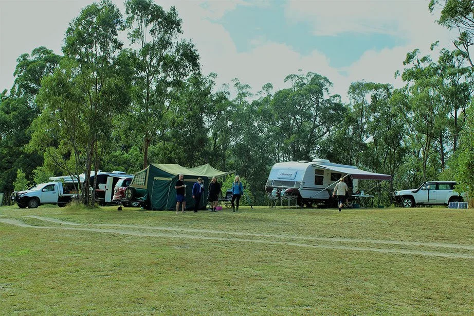 A group of people at a campsite with tents, parked cars, and a traveling trailer among trees in a grassy area.