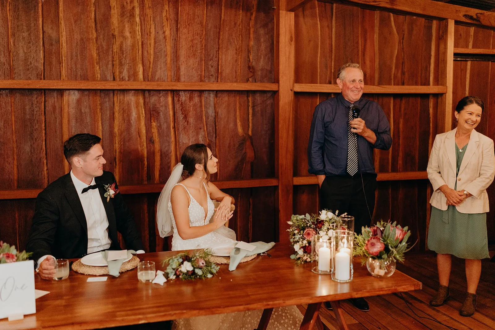 A wedding reception with a bride and groom seated at a wooden table, listening to a man giving a speech, with a woman standing nearby, in a rustic wooden venue decorated with flowers and candles.