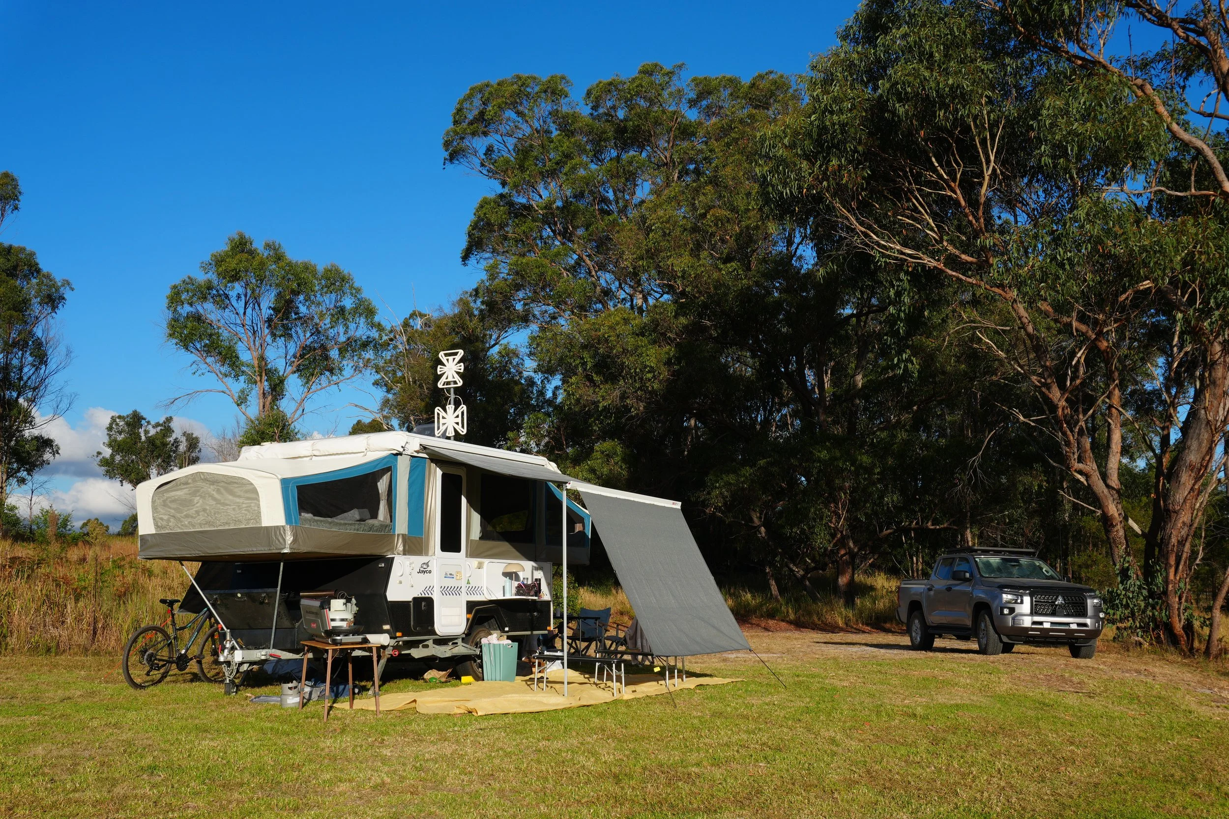 A campsite with a camper trailer and a roof tent set up in an open field, surrounded by trees under a bright blue sky.