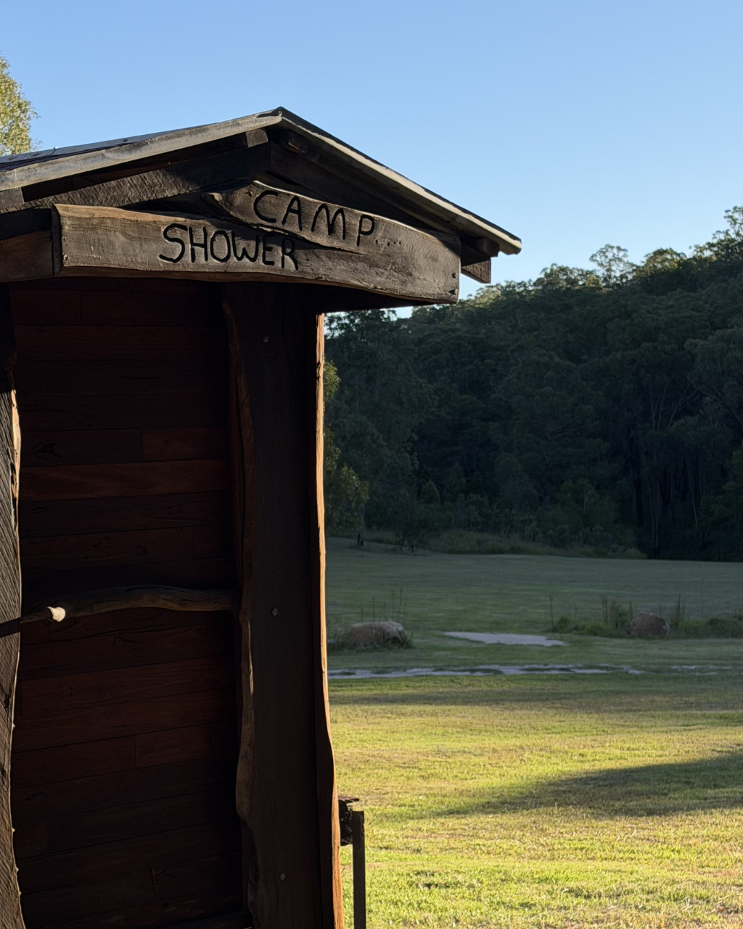 Wooden outdoor shower shelter with a sign that reads 'CAMP SHOWER'. Green field and trees in the background under a clear blue sky.