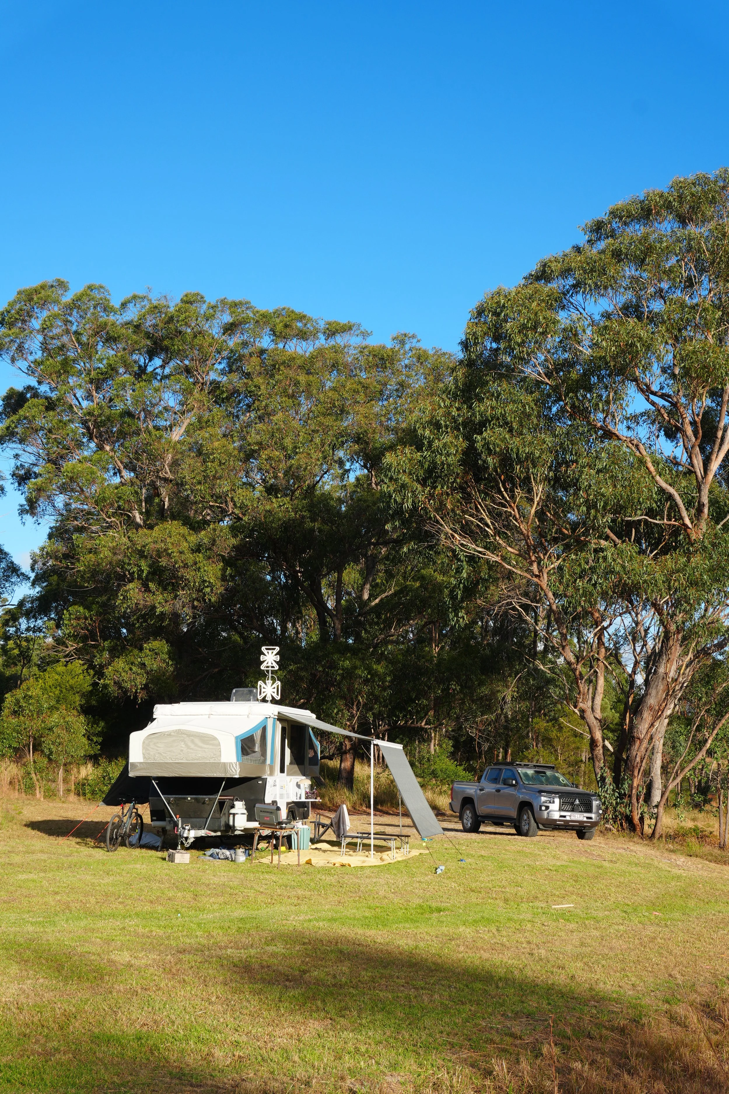 A campsite with a white camper trailer, a grey pickup truck, and outdoor furniture, set in a grassy area with tall trees and a bright blue sky.