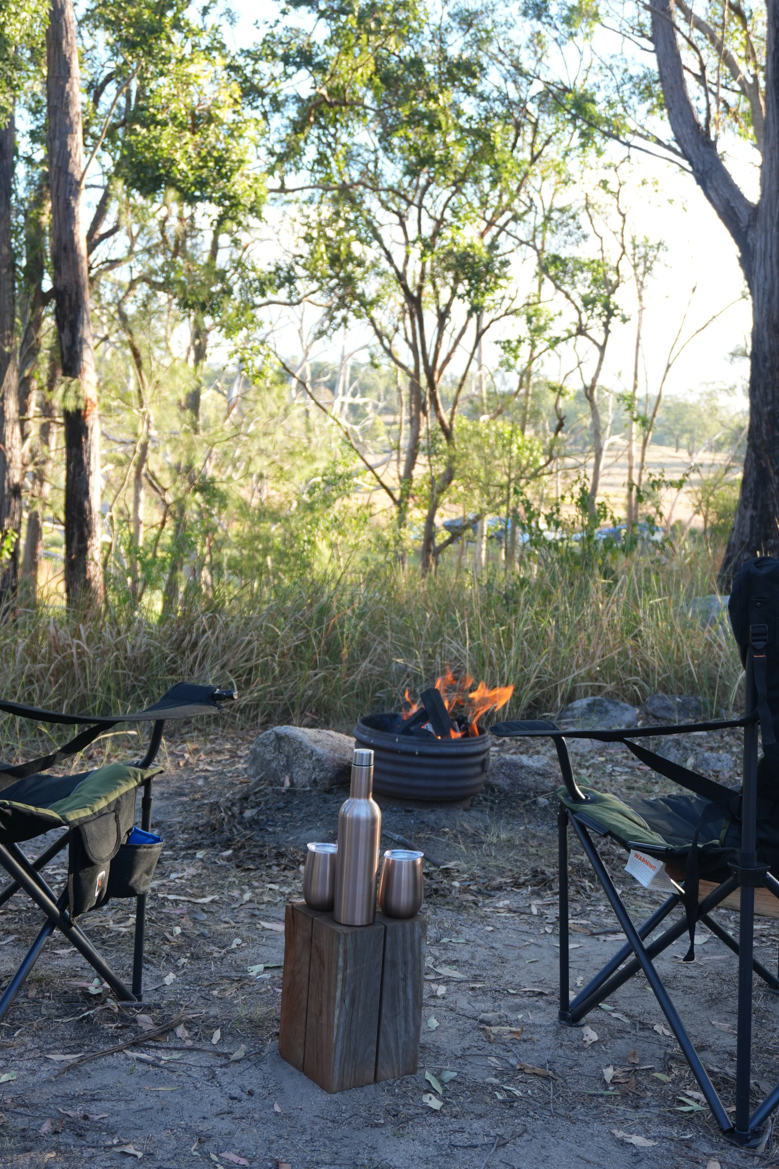 Camping scene with two chairs, a small wooden table holding three metal cups, a portable stove, and a small fire pit in a wooded area with trees and tall grass in the background.