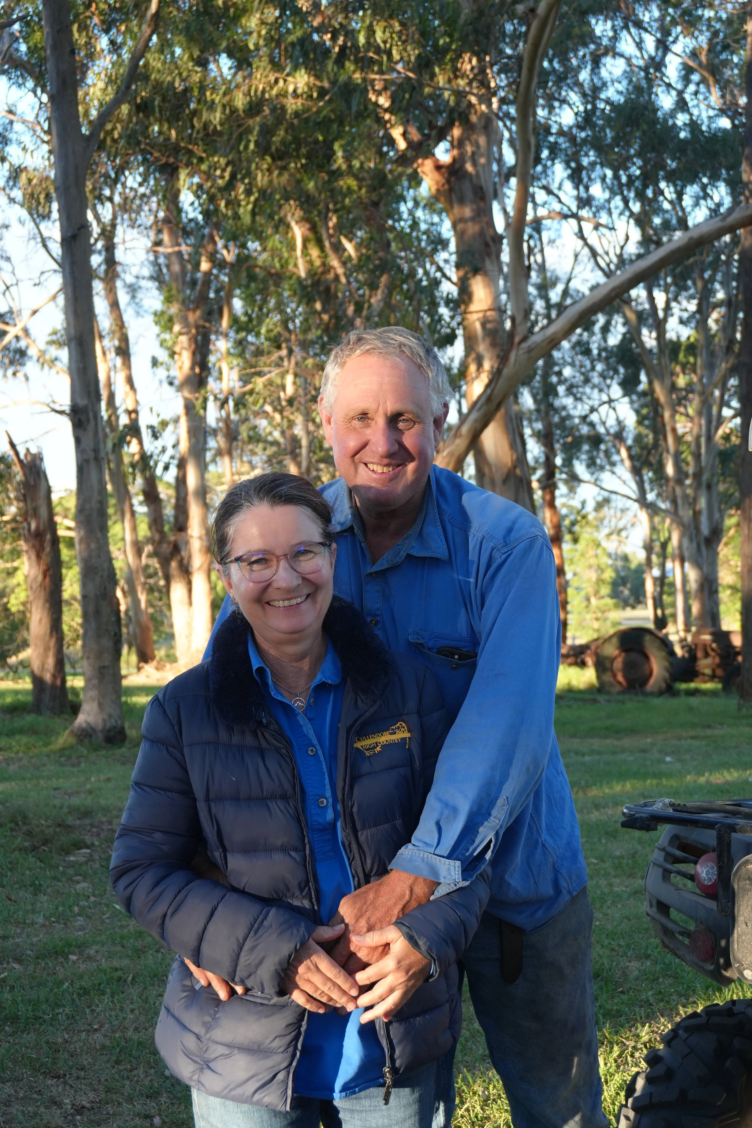 A smiling woman with glasses and dark hair, and a man, both outdoors among tall trees, standing close together and holding hands. The woman is wearing a navy blue jacket with a logo on it, and the man is wearing a blue shirt. The background includes green grass and trees, with a tractor partially visible on the right side.