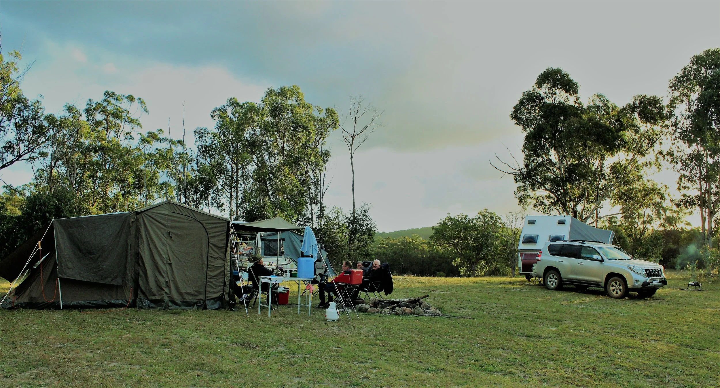 People camping outdoors with tents, a camper, and a car in a grassy field surrounded by trees, during late afternoon or early evening.